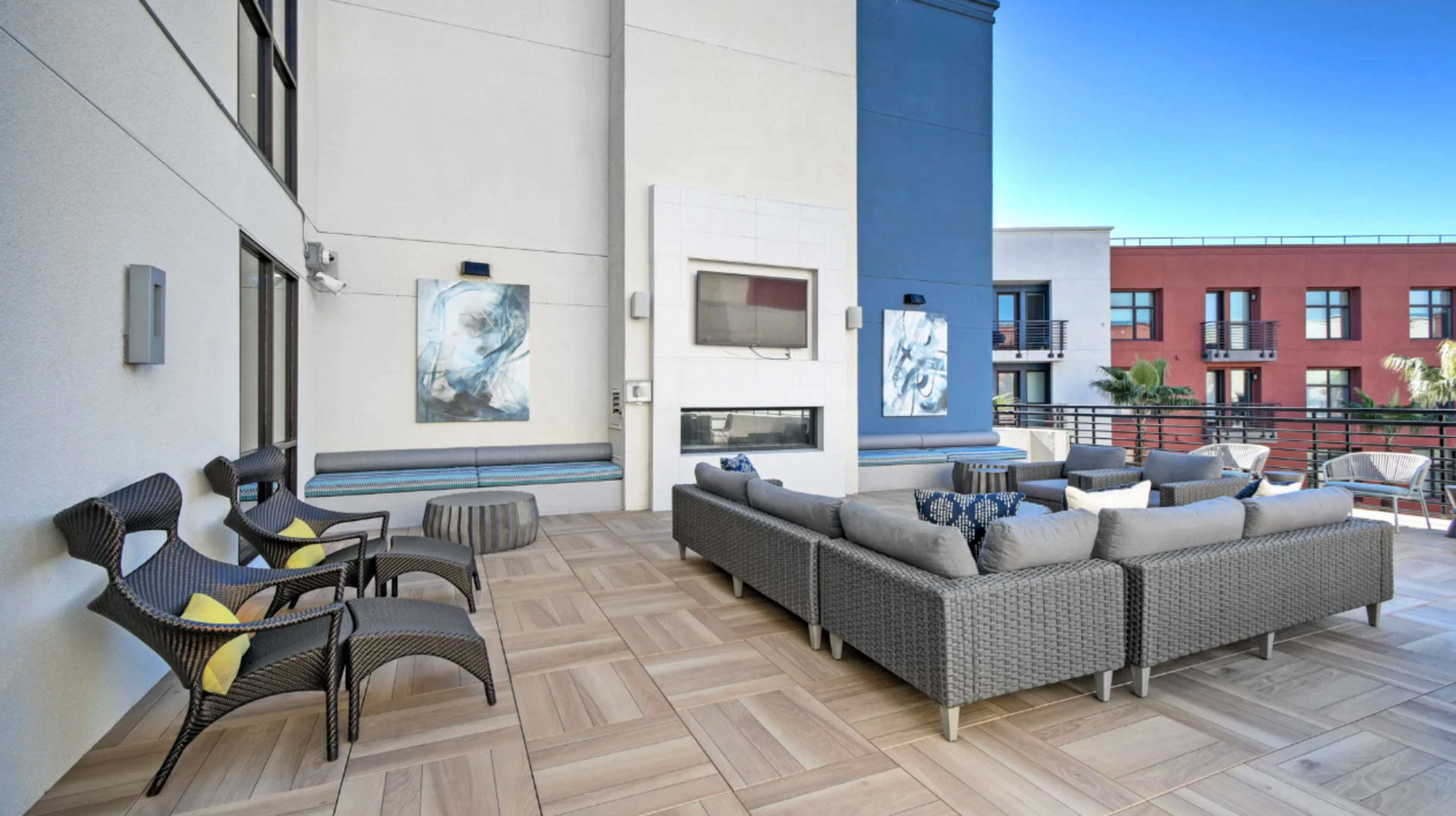 A modern outdoor patio area with a sectional sofa, armchairs, and artwork on the walls, overlooking a sunny urban environment.