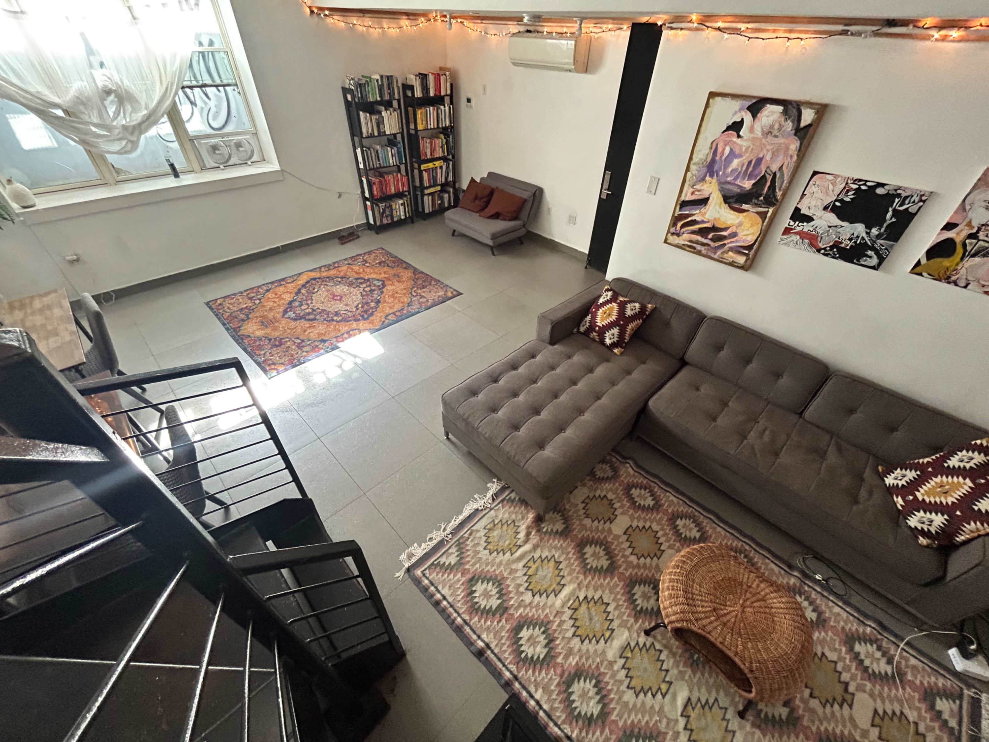 A living room features a sectional couch, a small table, several rugs, and bookshelves along the walls, with decorative artwork displayed.