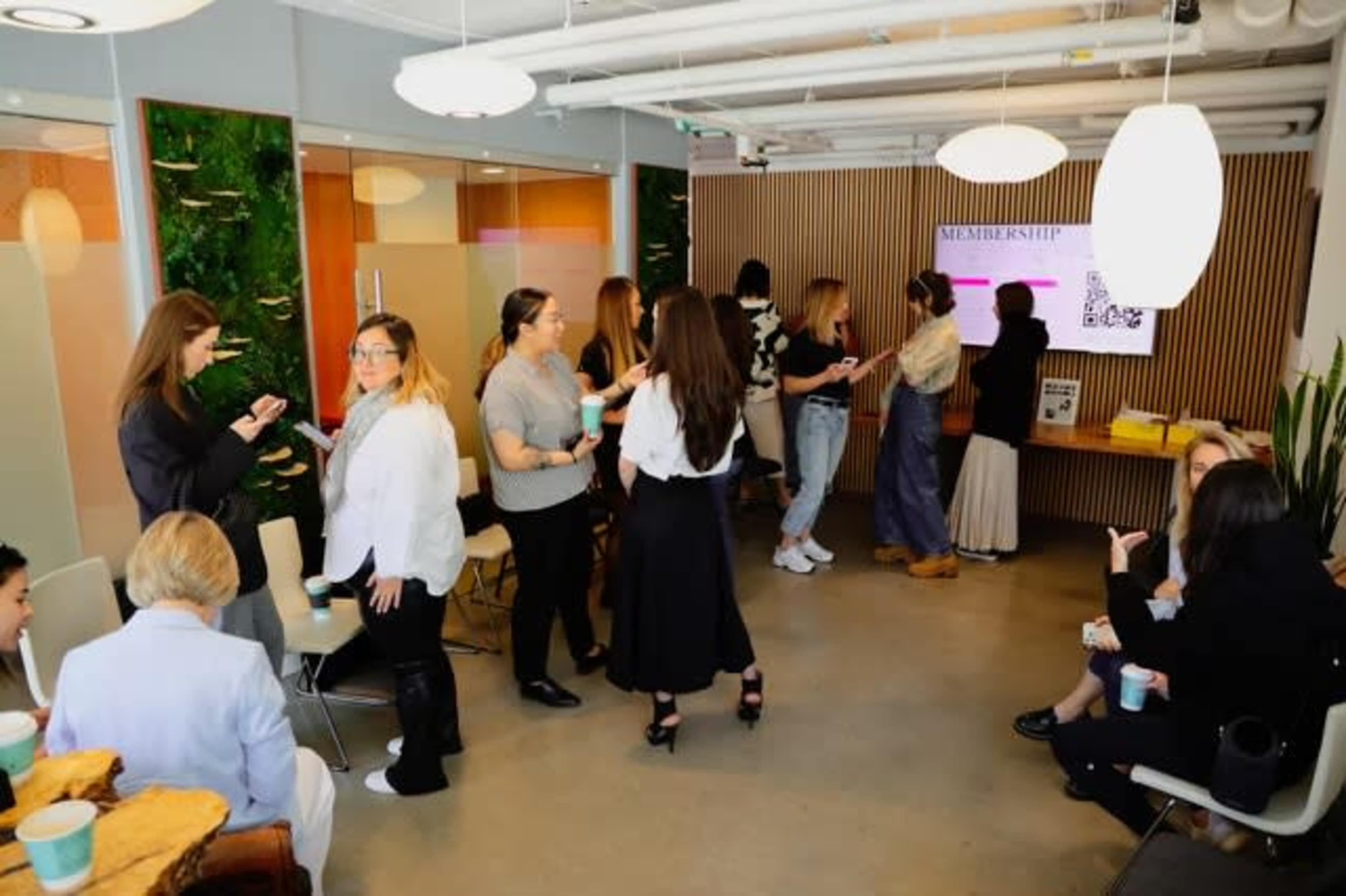 A group of women socialize in a modern office space featuring a digital display and green wall elements.
