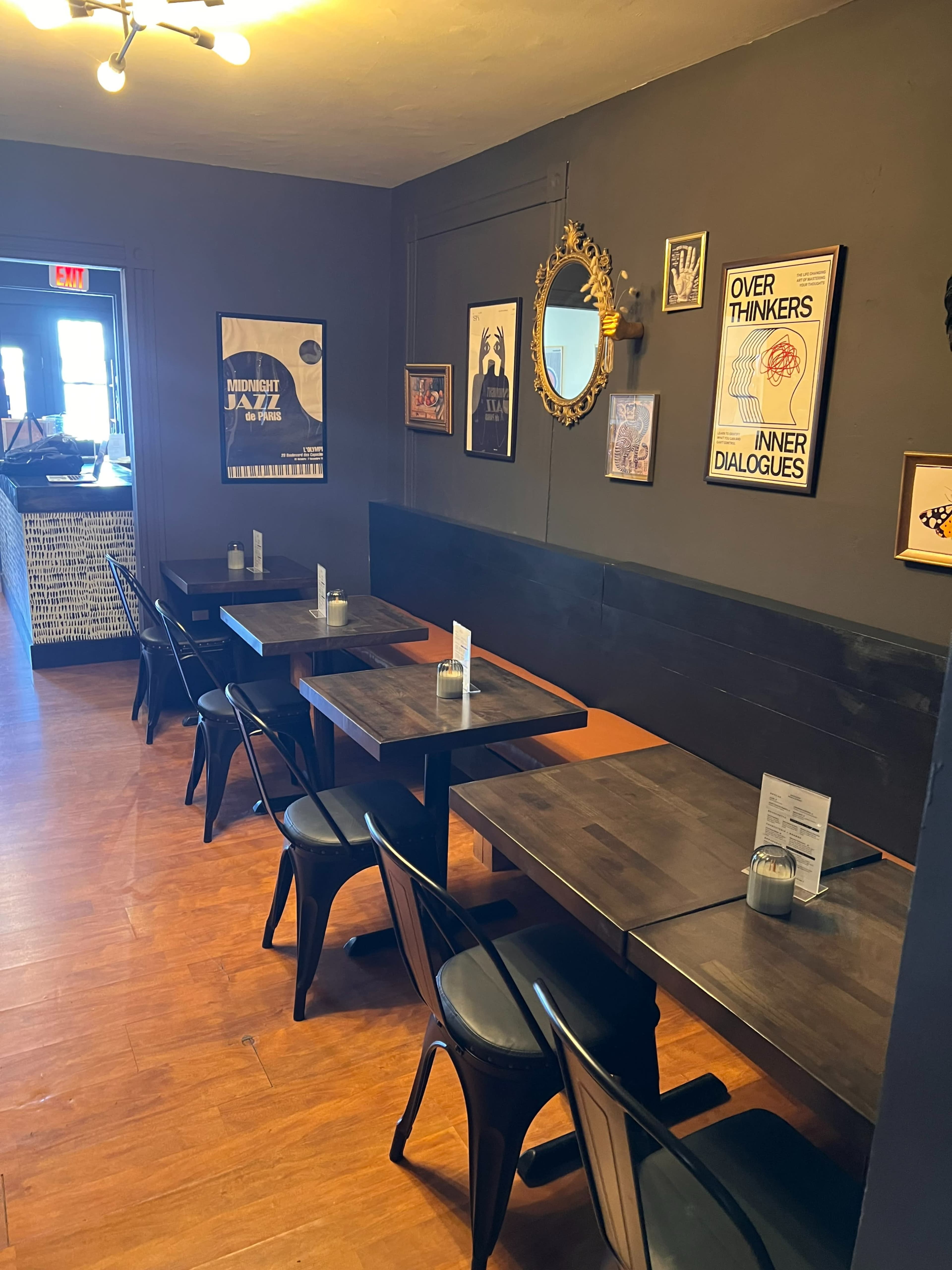 The image shows a narrow restaurant interior with black tables and chairs arranged along a wall, adorned with various framed posters and a decorative mirror.