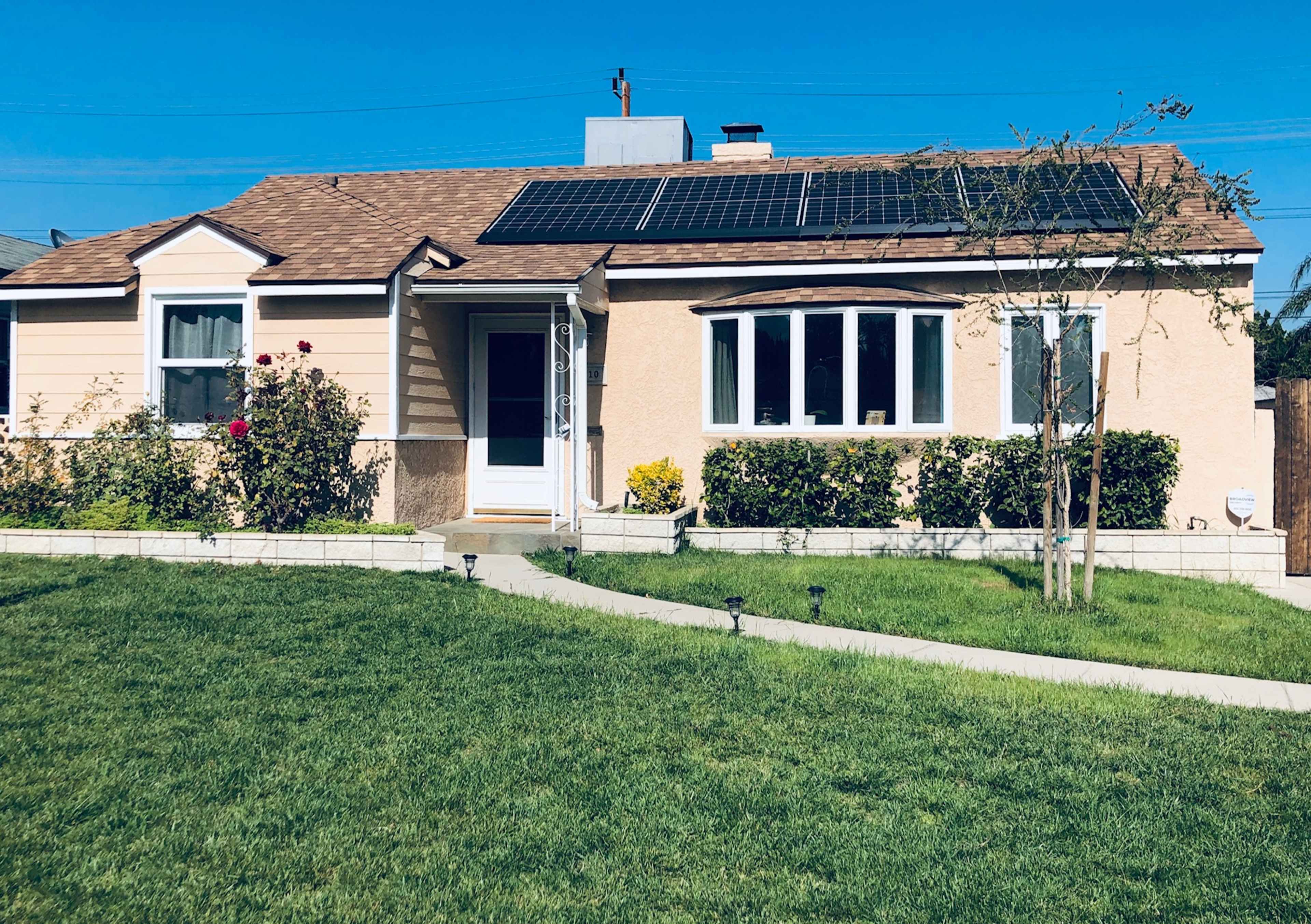 A single-story house with solar panels on the roof is surrounded by a green lawn and flower beds.