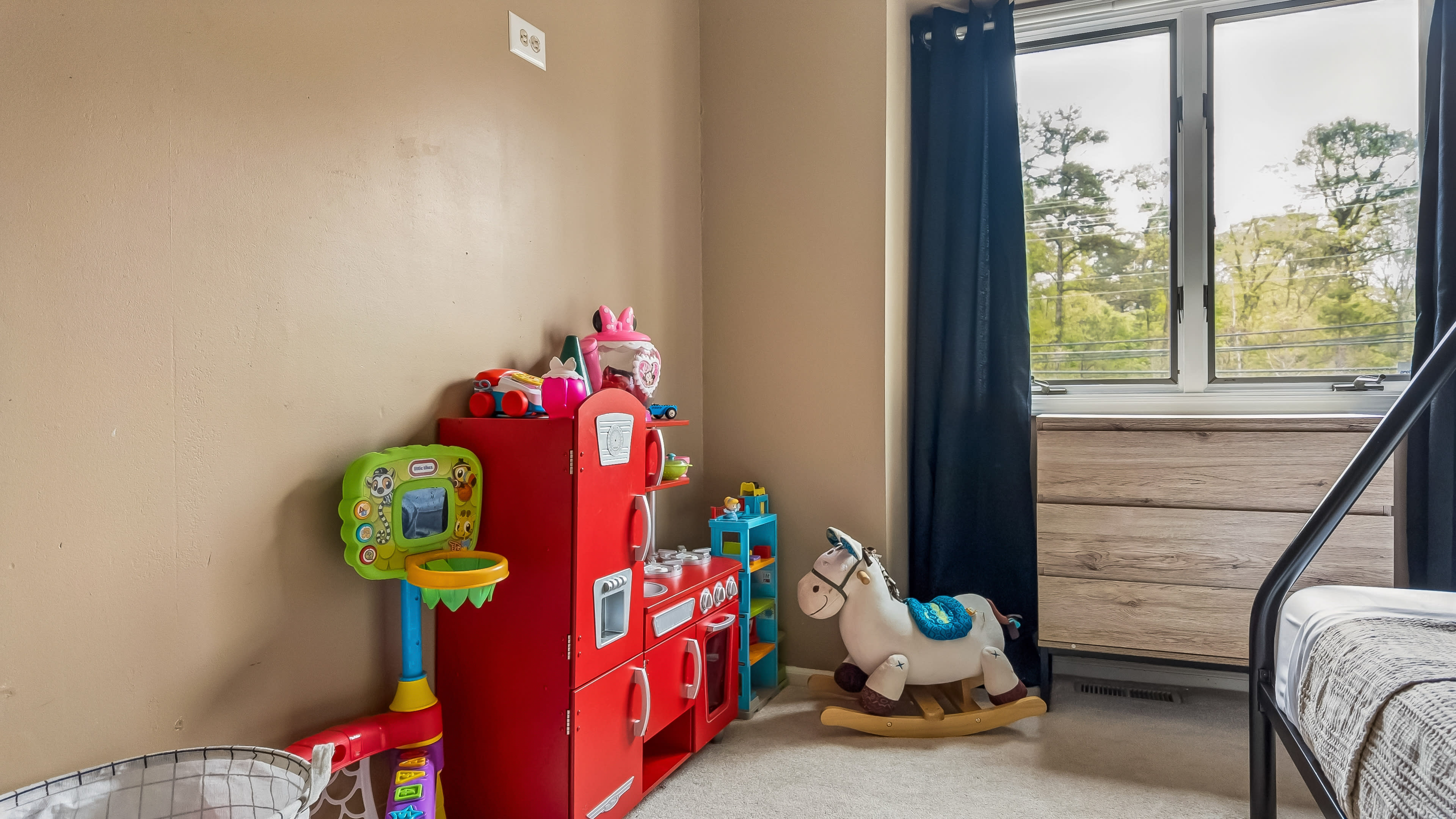 The image shows a child's bedroom with a red toy kitchen set, a rocking horse, and a dresser near a window with dark curtains.