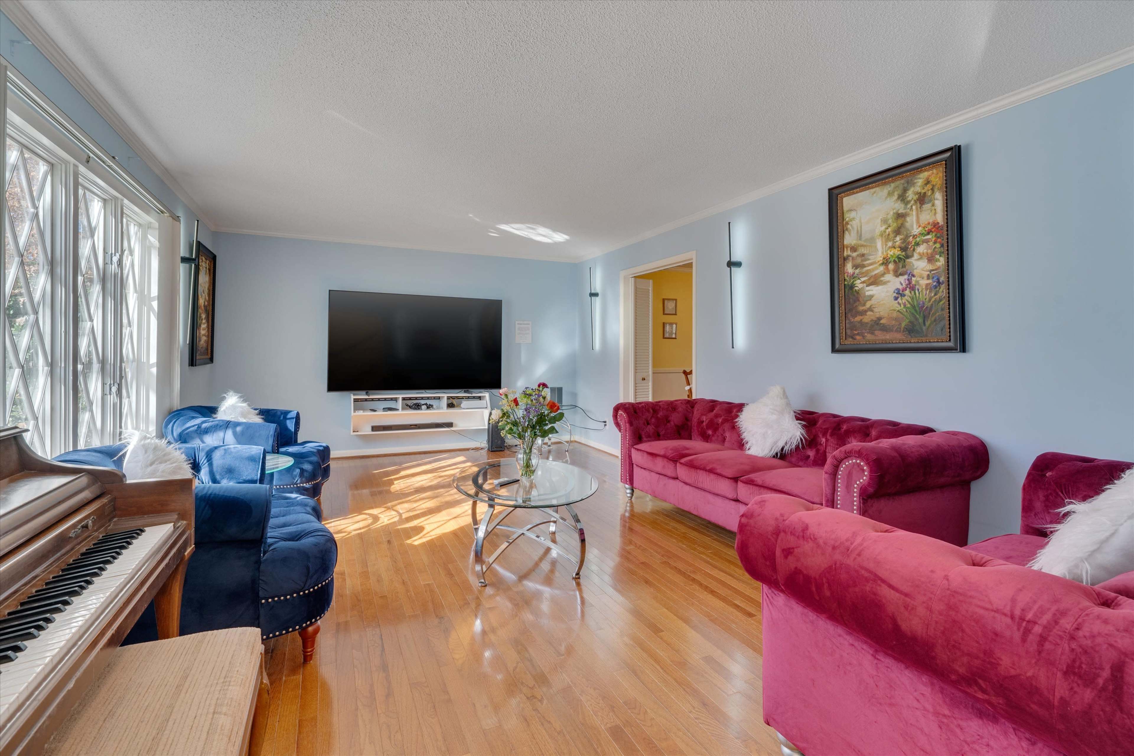 The living room features a piano, a glass coffee table, and seating arrangements with blue and burgundy sofas against light blue walls.