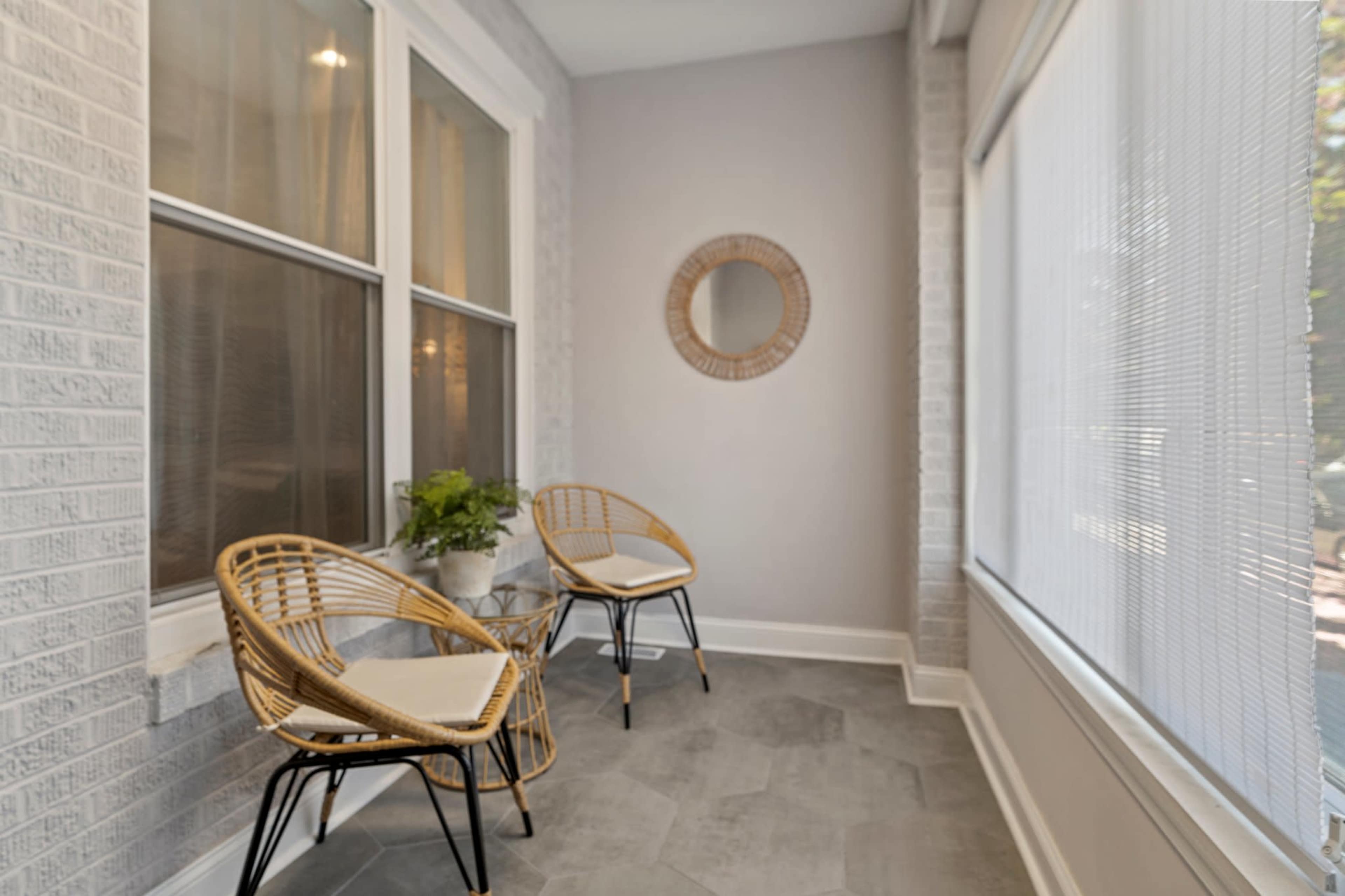 A small sunroom featuring two rattan chairs, a glass table, and a round mirror, with large windows allowing natural light to enter.