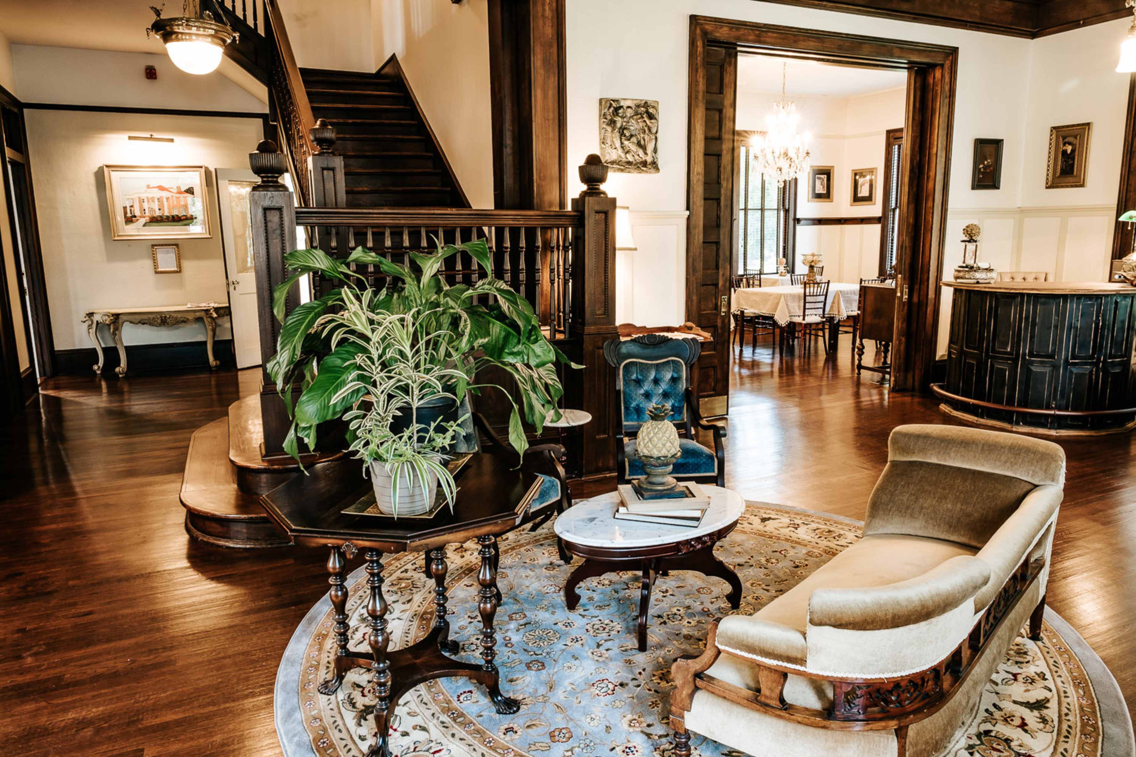 A vintage-style foyer with a staircase, a seating area featuring a plant and decorative furniture, and a dining area visible in the background.