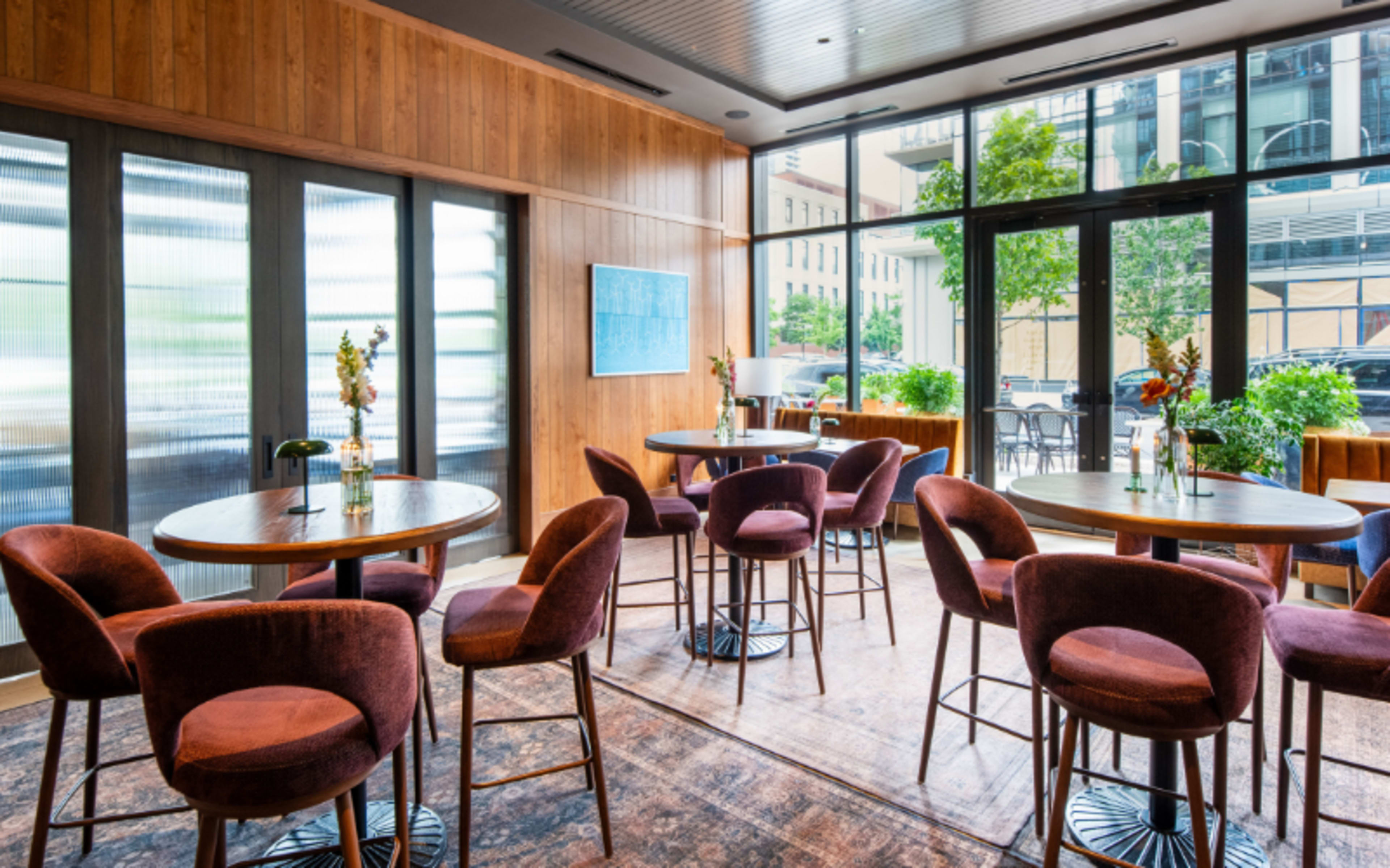 The image shows a modern dining area with round tables, high chairs, and large windows that overlook an outdoor space.