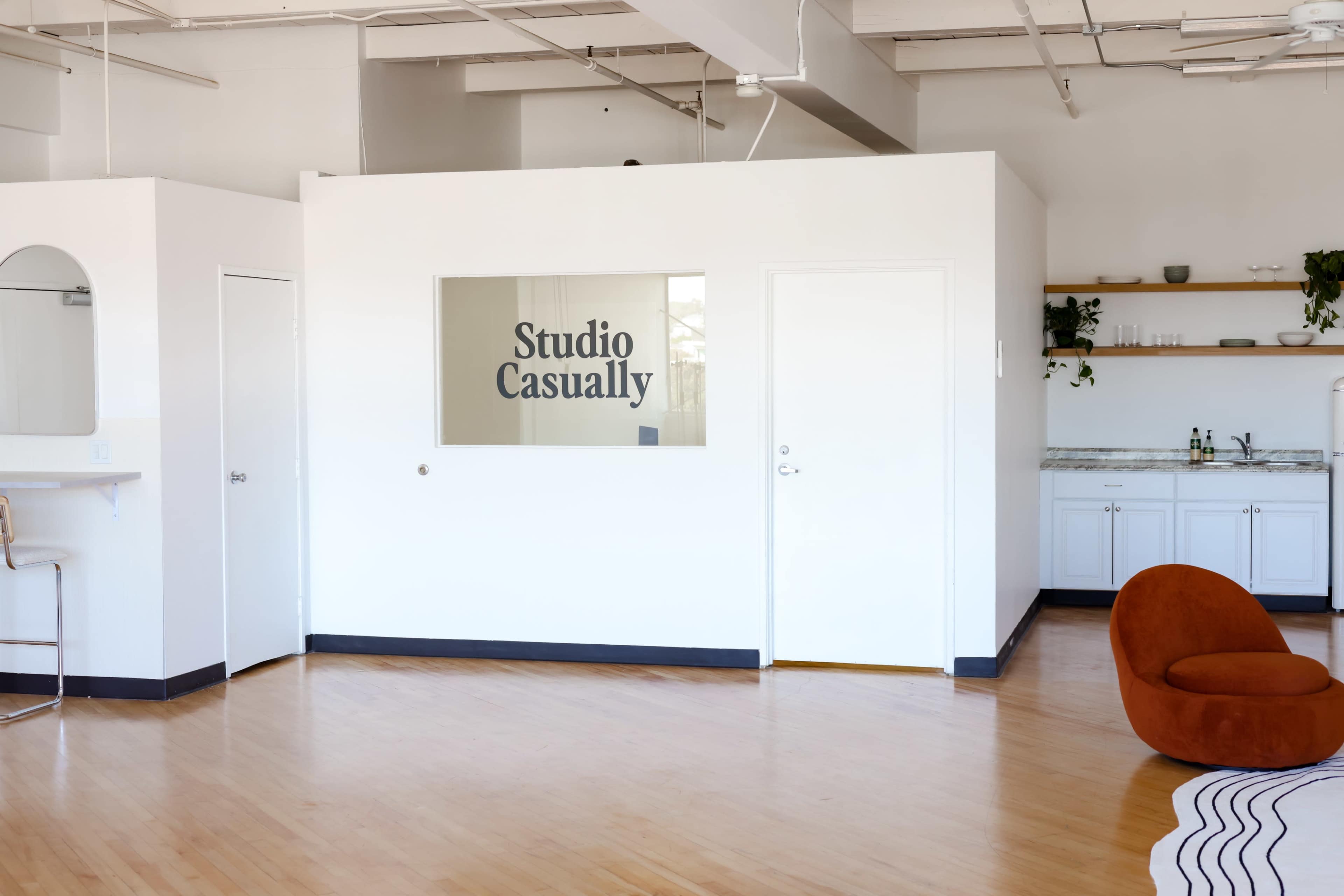 A bright, open space featuring a wall with the words "Studio Casually" displayed in a glass panel, alongside a cozy brown chair and a kitchenette area with white cabinetry.