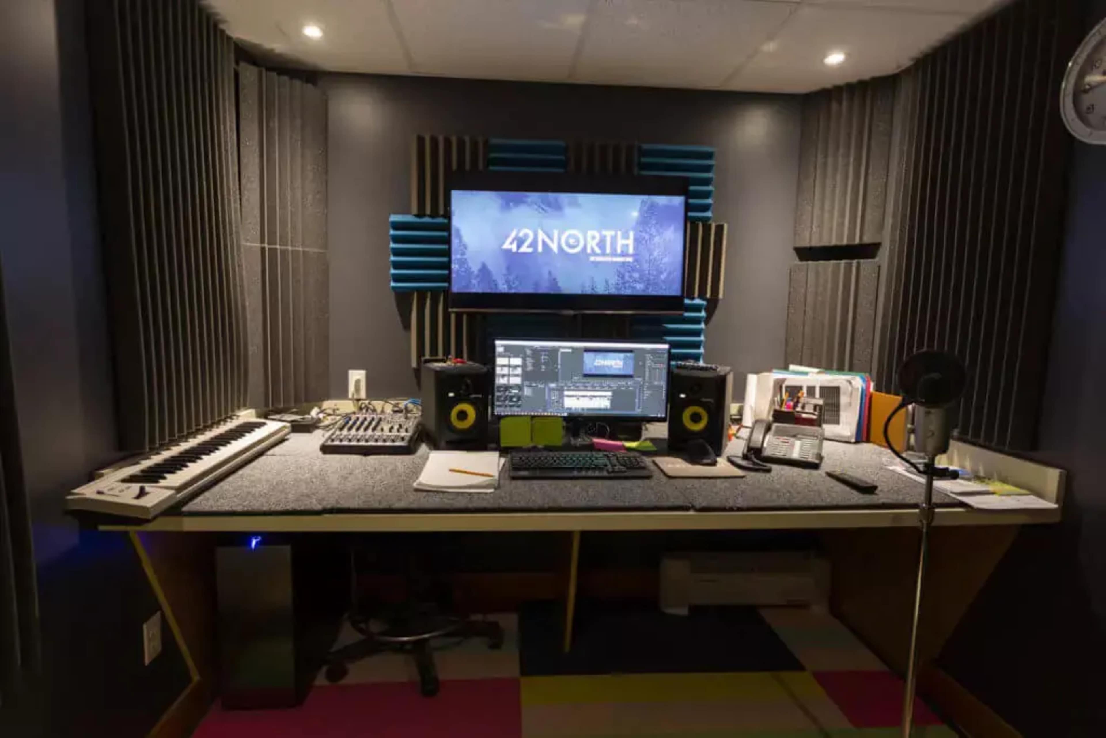 The image shows a music production studio with a large screen displaying "42 NORTH," a keyboard, audio equipment, and an organized workspace.