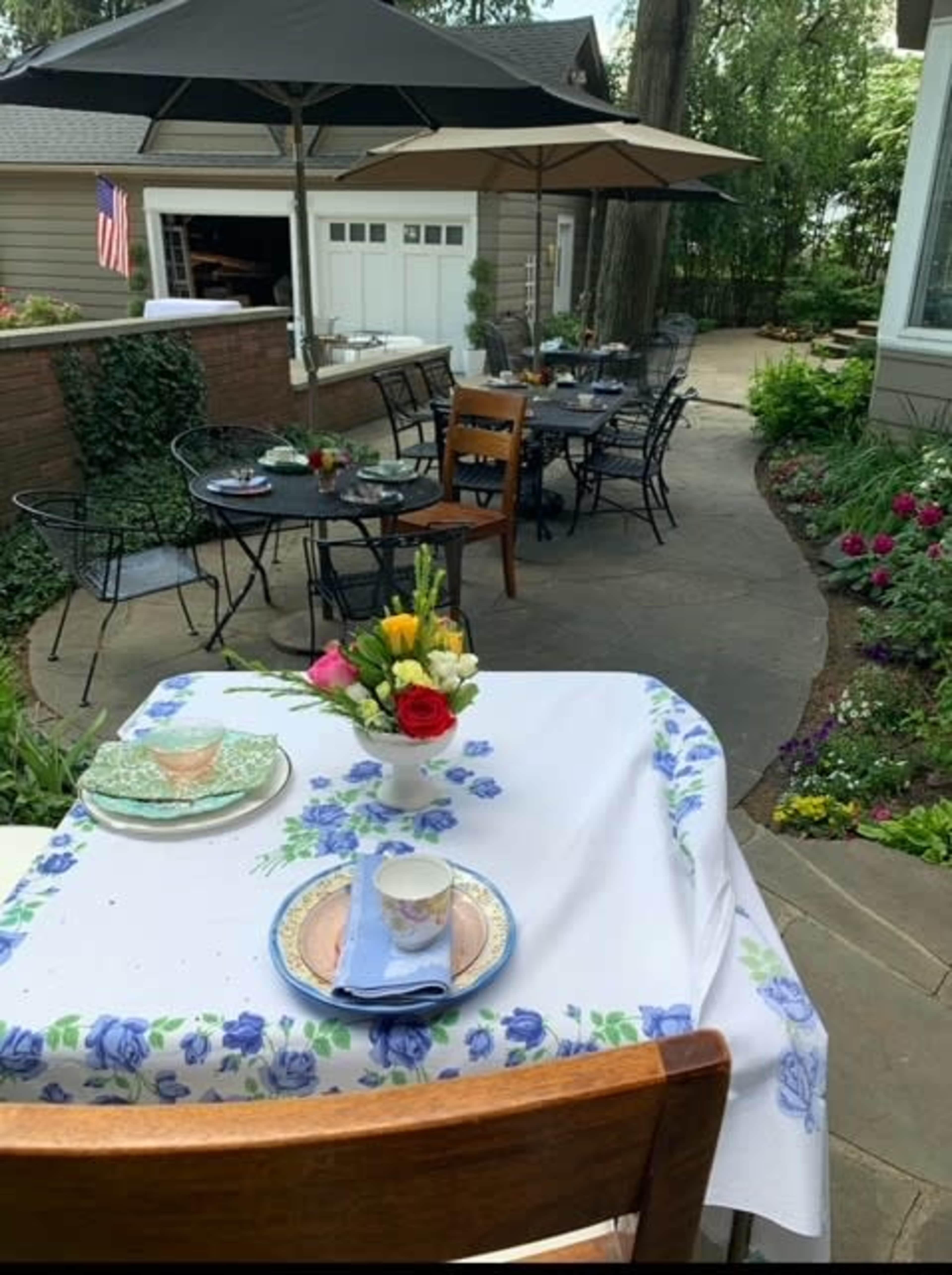 An outdoor dining area features tables set with floral tablecloths and chairs, surrounded by lush greenery and blooming flowers.