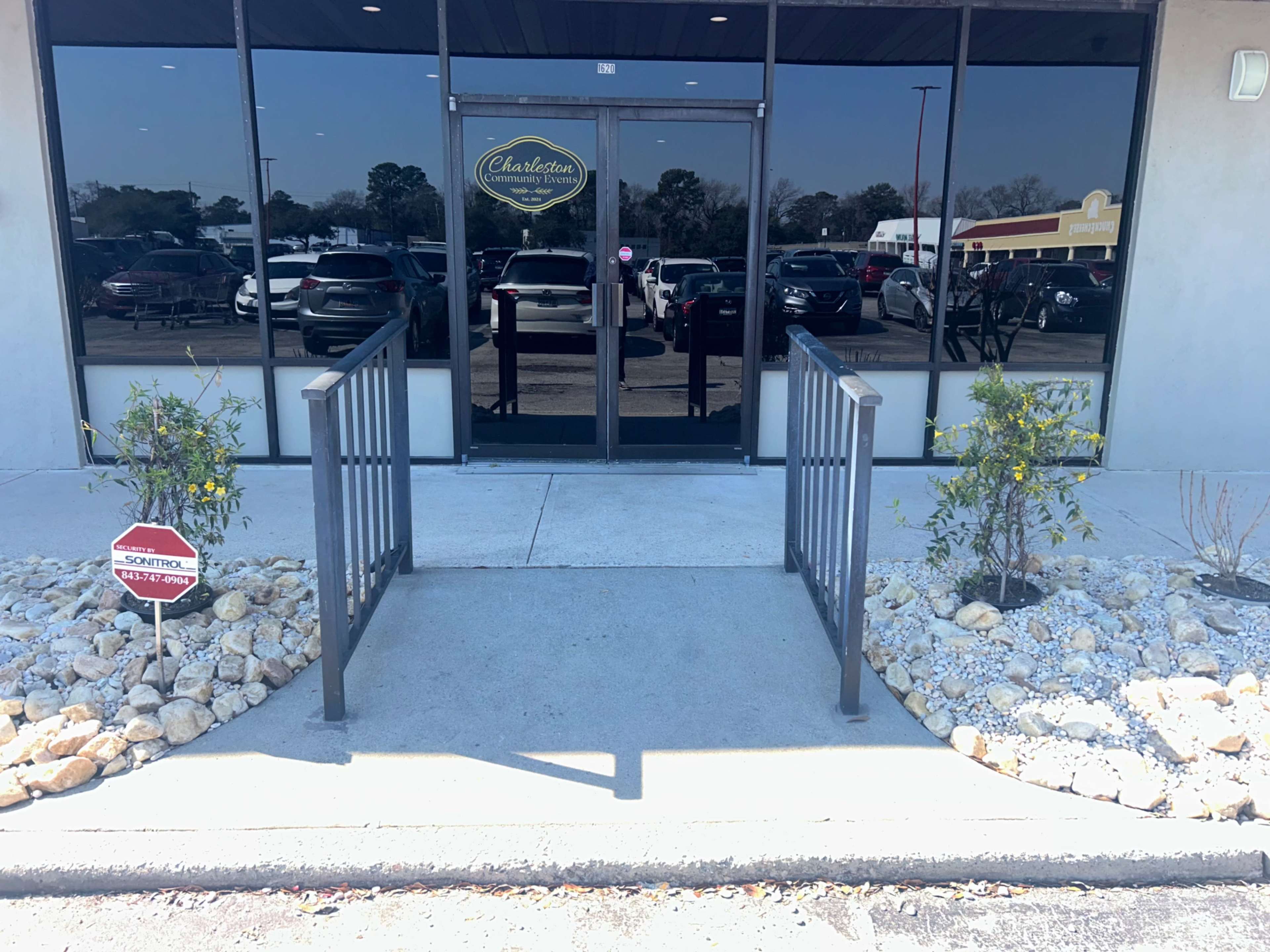The image shows a building entrance with a glass door flanked by planters, a small ramp, and a signpost.