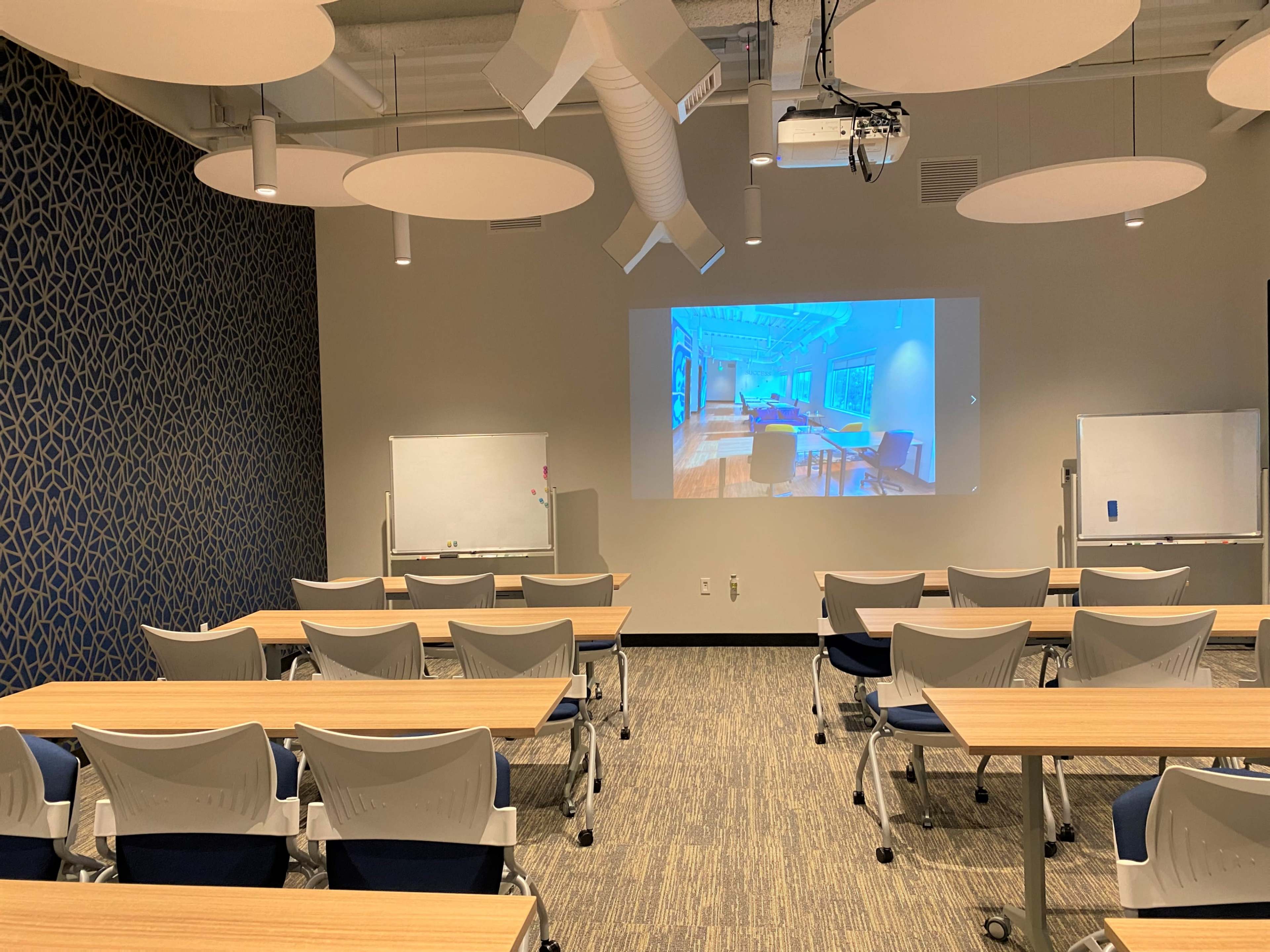 A modern meeting room with rows of tables and chairs, a projection screen displaying a blue-toned interior design, and whiteboards on either side.