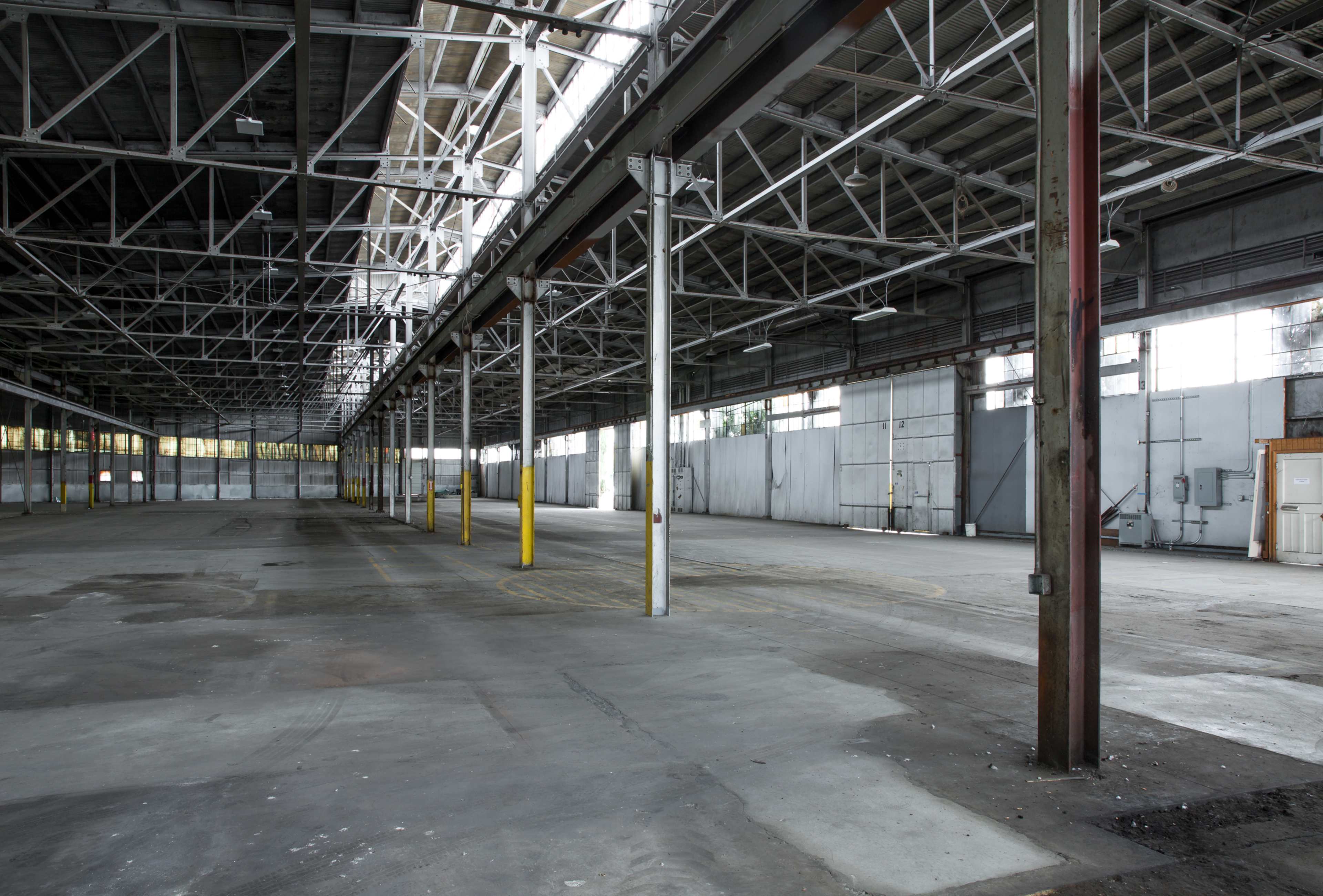 A large, empty warehouse interior with high ceilings and steel support beams, featuring concrete flooring and sparse lighting.