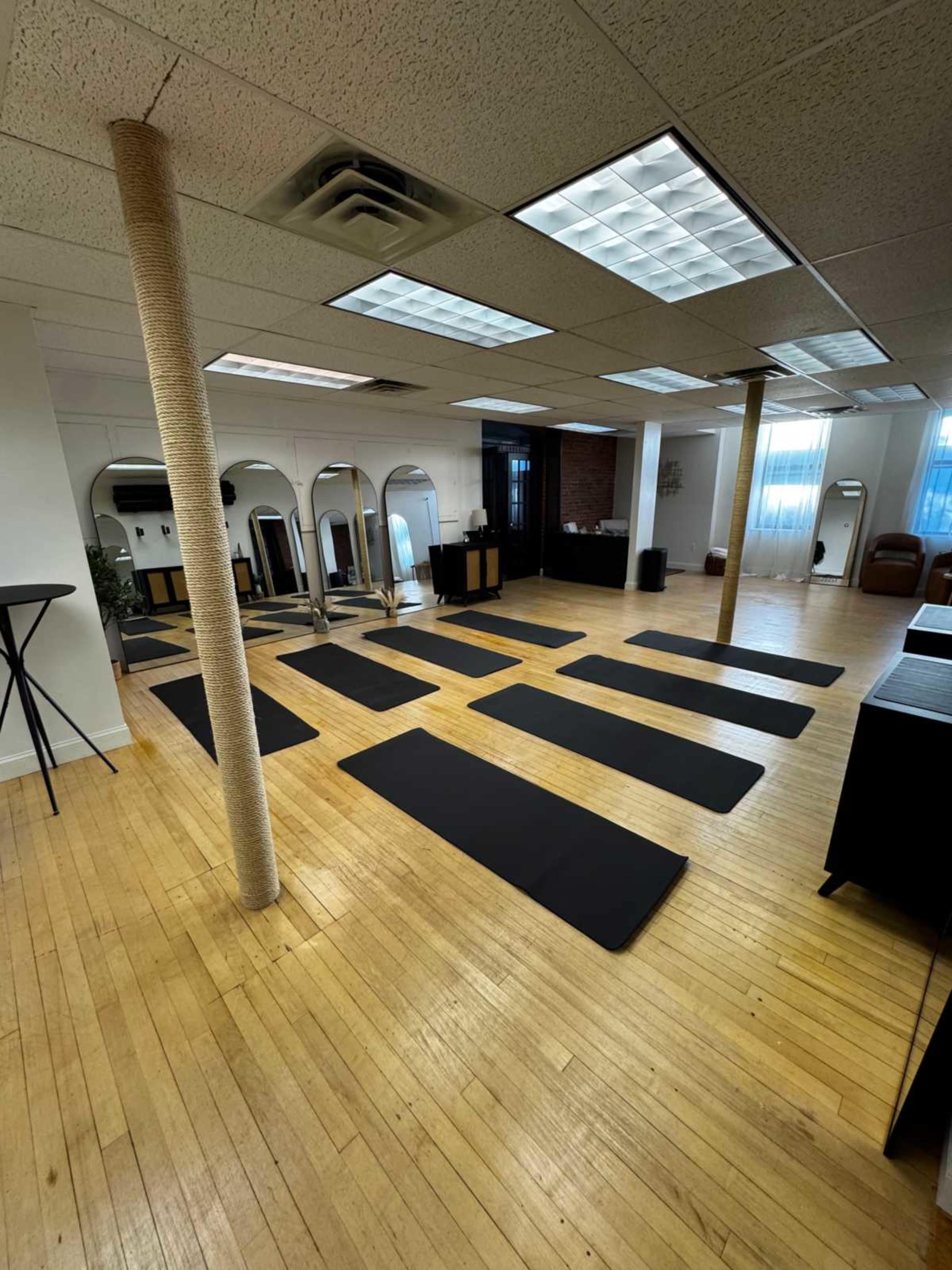 The image shows a spacious, well-lit studio with multiple yoga mats arranged neatly on a wooden floor.