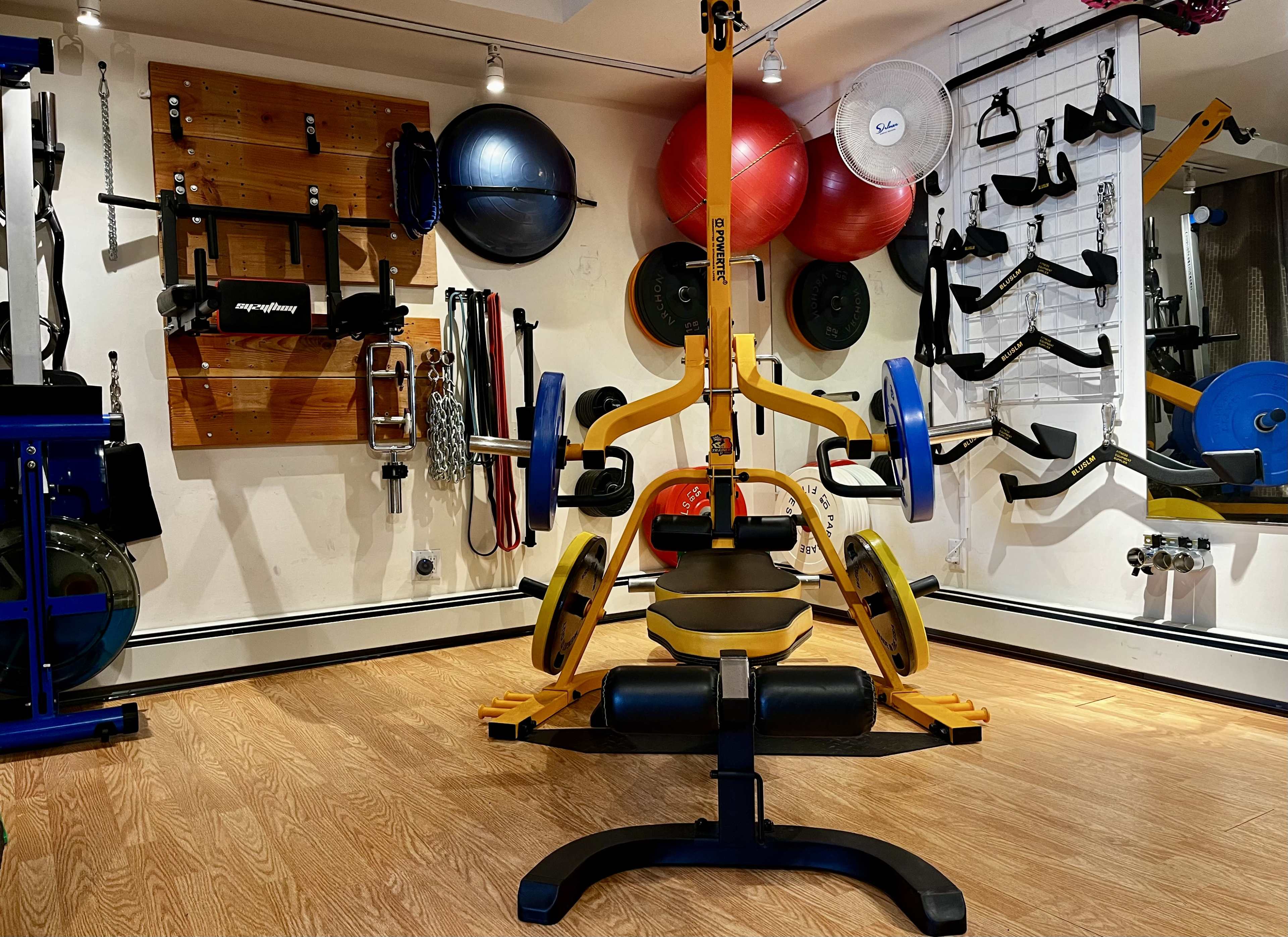 A well-equipped home gym featuring a yellow weight bench with barbells, various exercise accessories, and colorful exercise balls against a wooden and white wall.