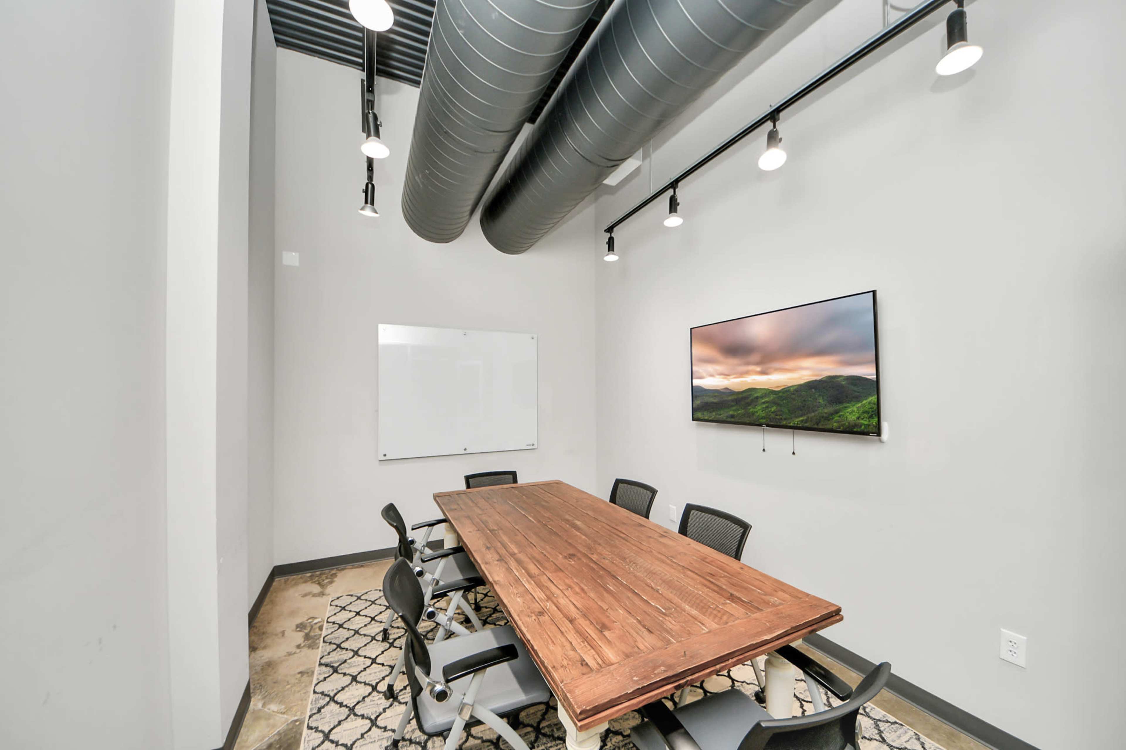Aboardroom features a long wooden table with eight chairs, a large wall-mounted television, a whiteboard, and exposed ductwork on the ceiling.