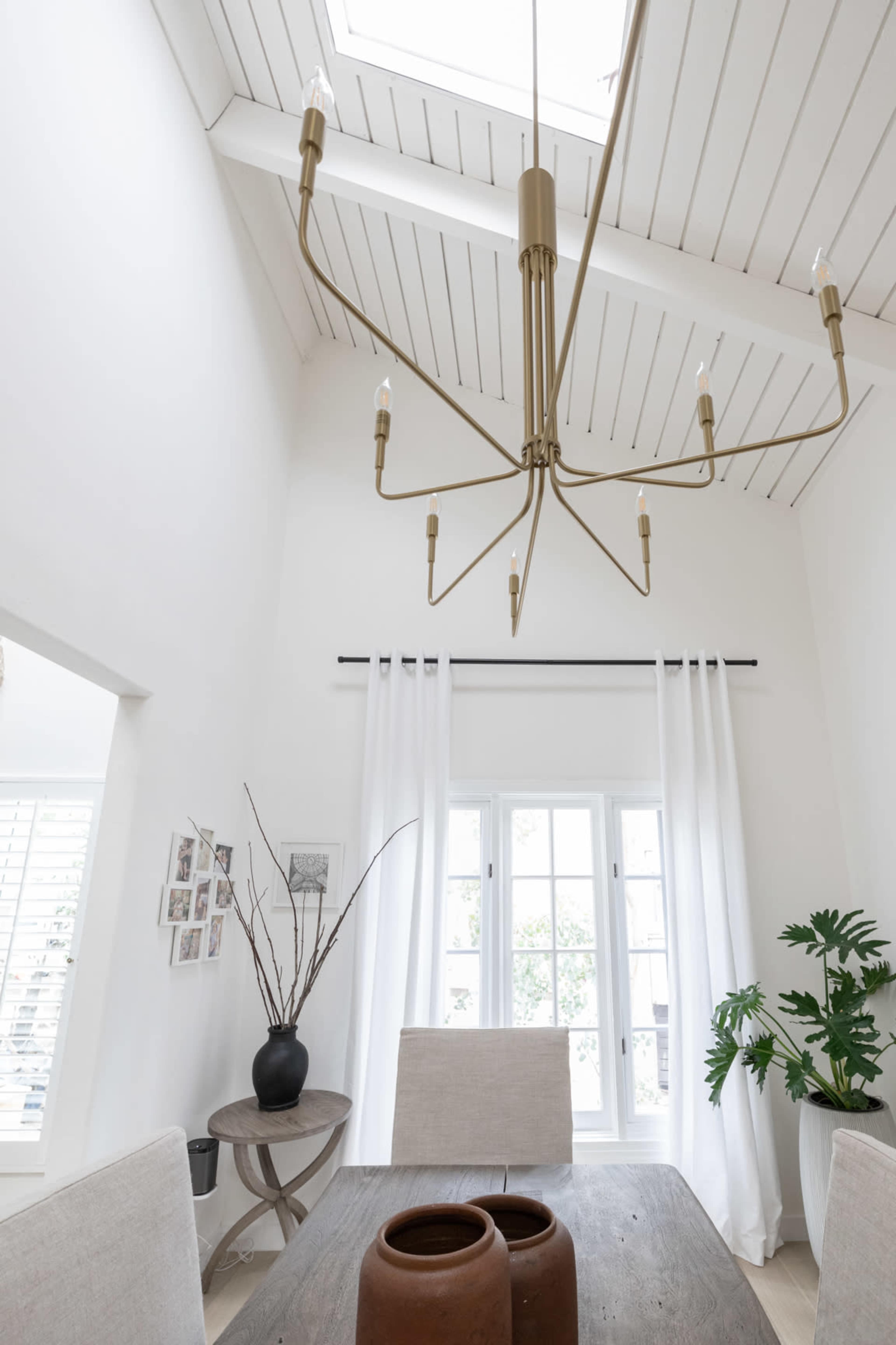 A bright dining area with a large chandelier hanging from a vaulted ceiling, a wooden table, and large windows draped with white curtains.