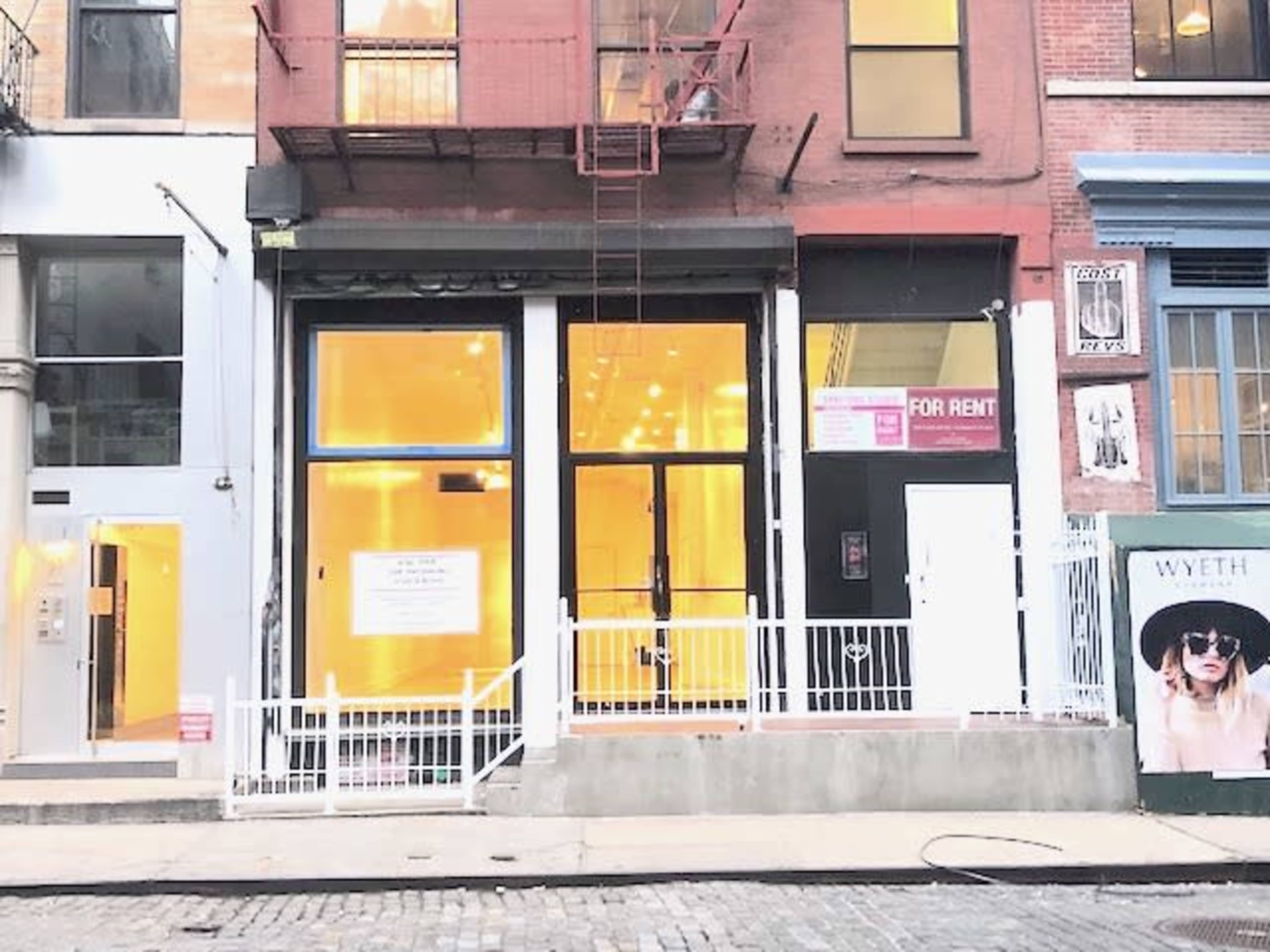 A storefront with large windows and a "For Rent" sign, flanked by two other commercial spaces, on a cobblestone street.