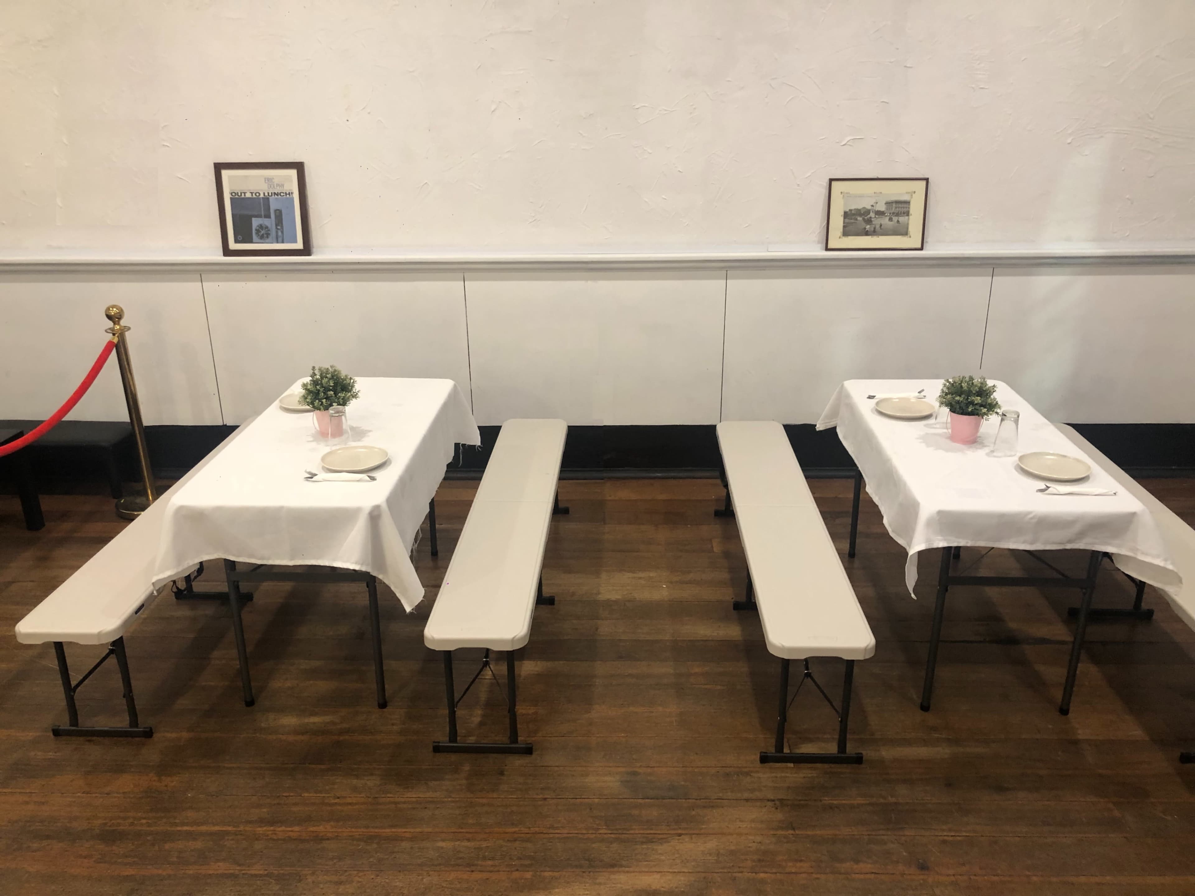 Two long tables with white tablecloths and centerpieces are set on a wooden floor, flanked by benches and framed pictures on the wall.