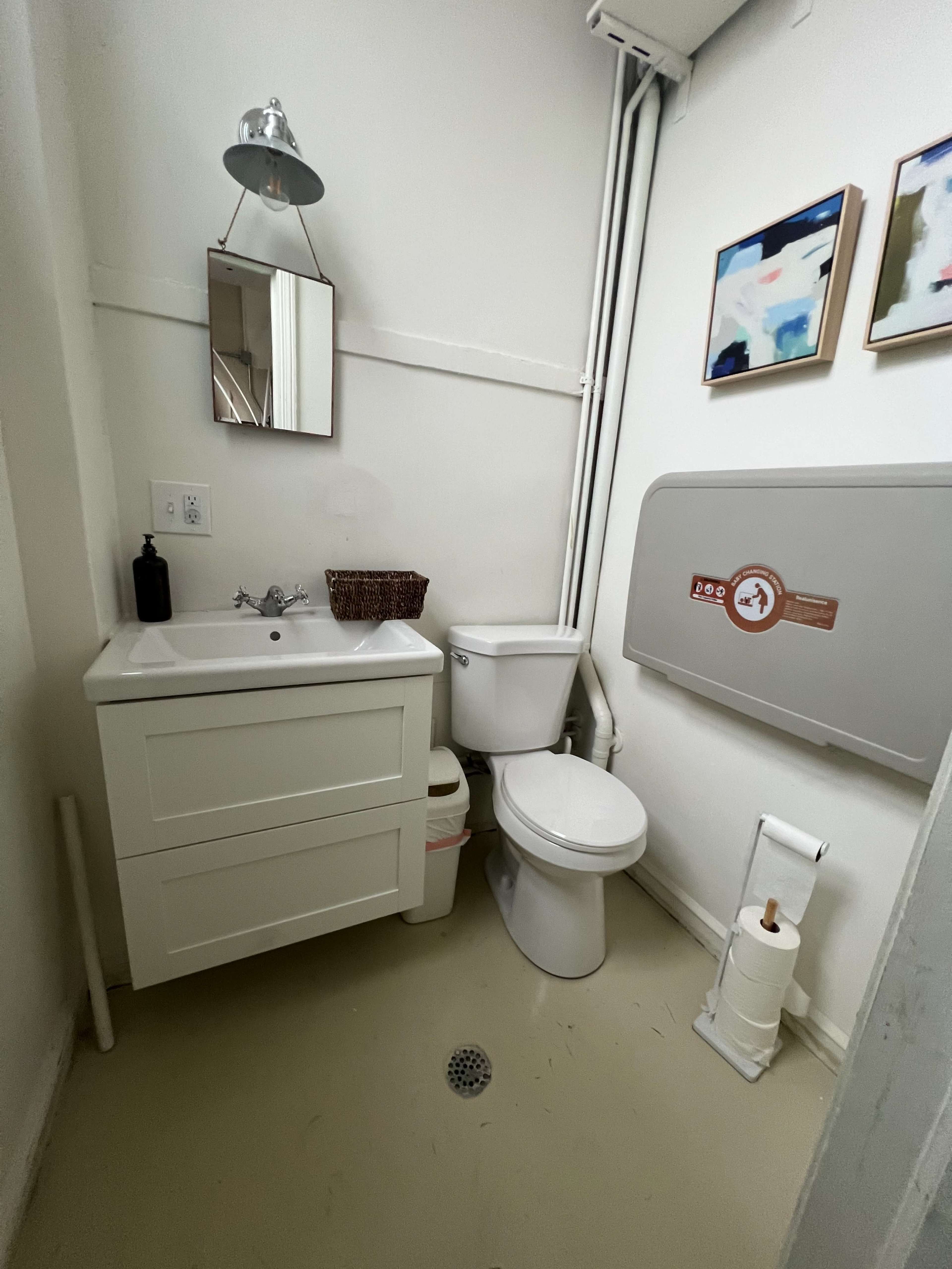 The image shows a small bathroom with a white sink and cabinet, a toilet, a wall-mounted mirror, and two framed artworks.