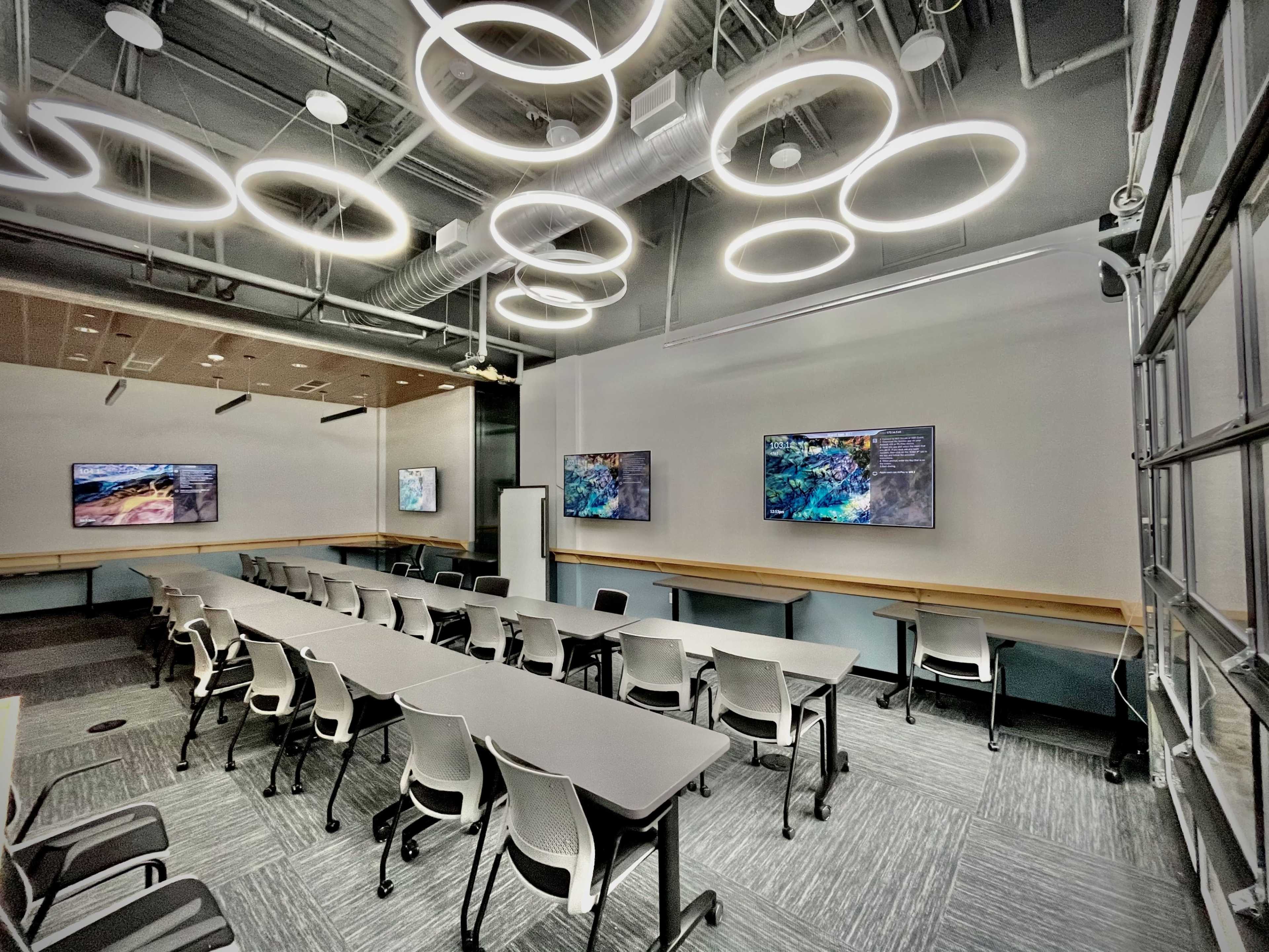 A modern classroom features circular light fixtures and two mounted televisions on the walls, with rows of tables and chairs arranged for instruction.