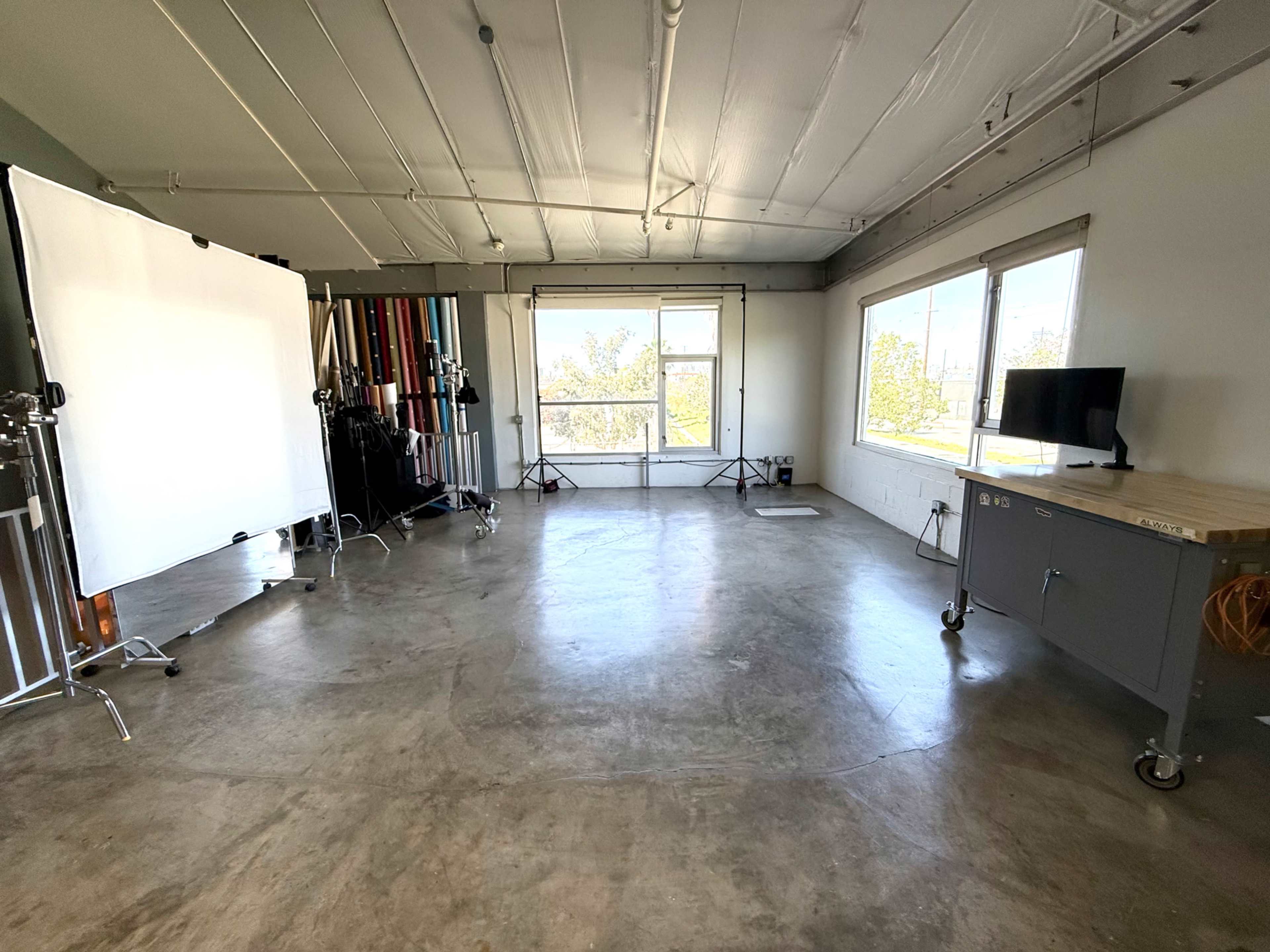 The image shows a spacious studio with a concrete floor, large windows, a digital screen on a cart, and lighting equipment positioned along one side.