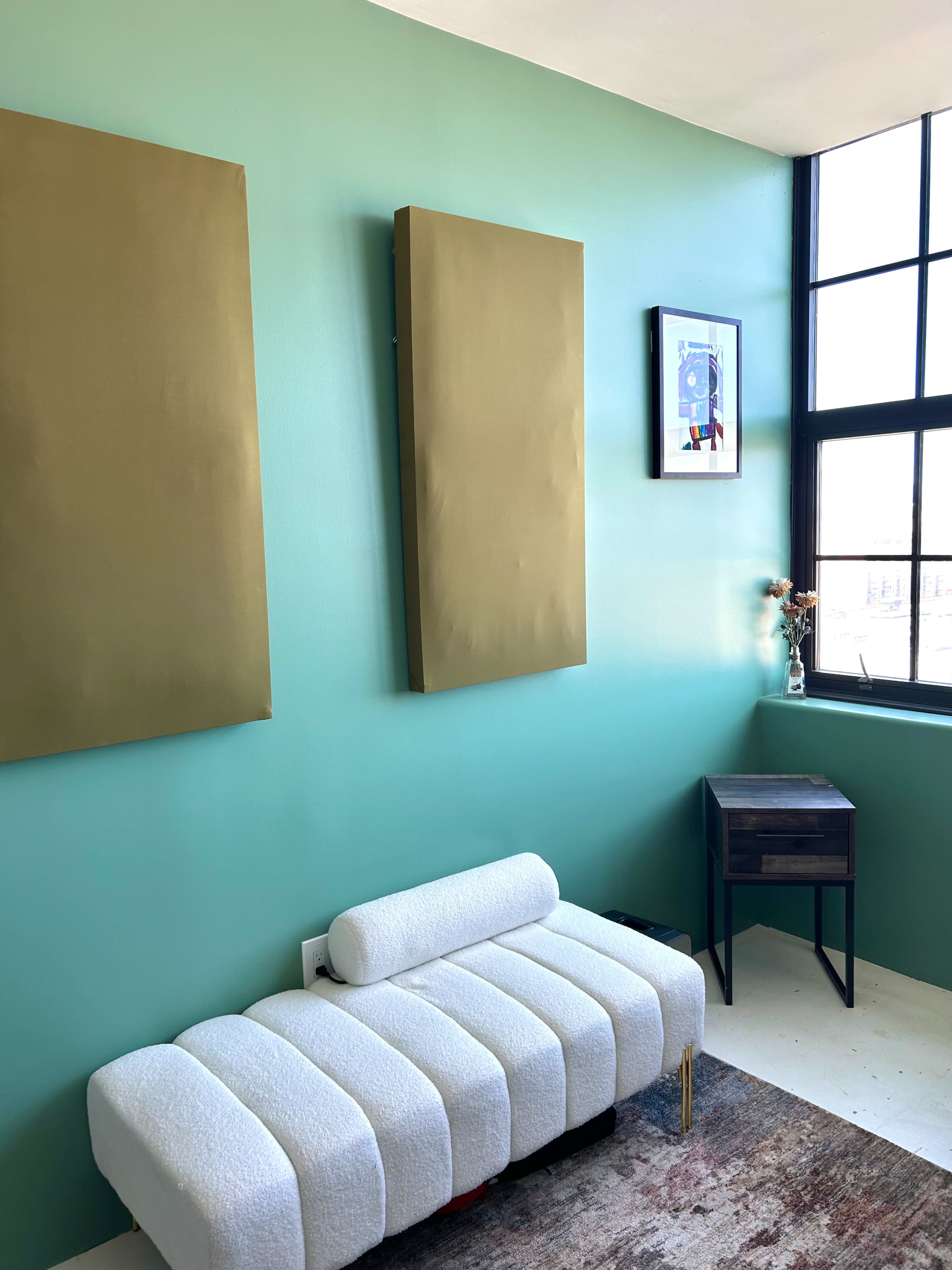 The image shows a room with turquoise walls, a white upholstered bench, two large gold artworks, and a small dark wooden side table beside a window.