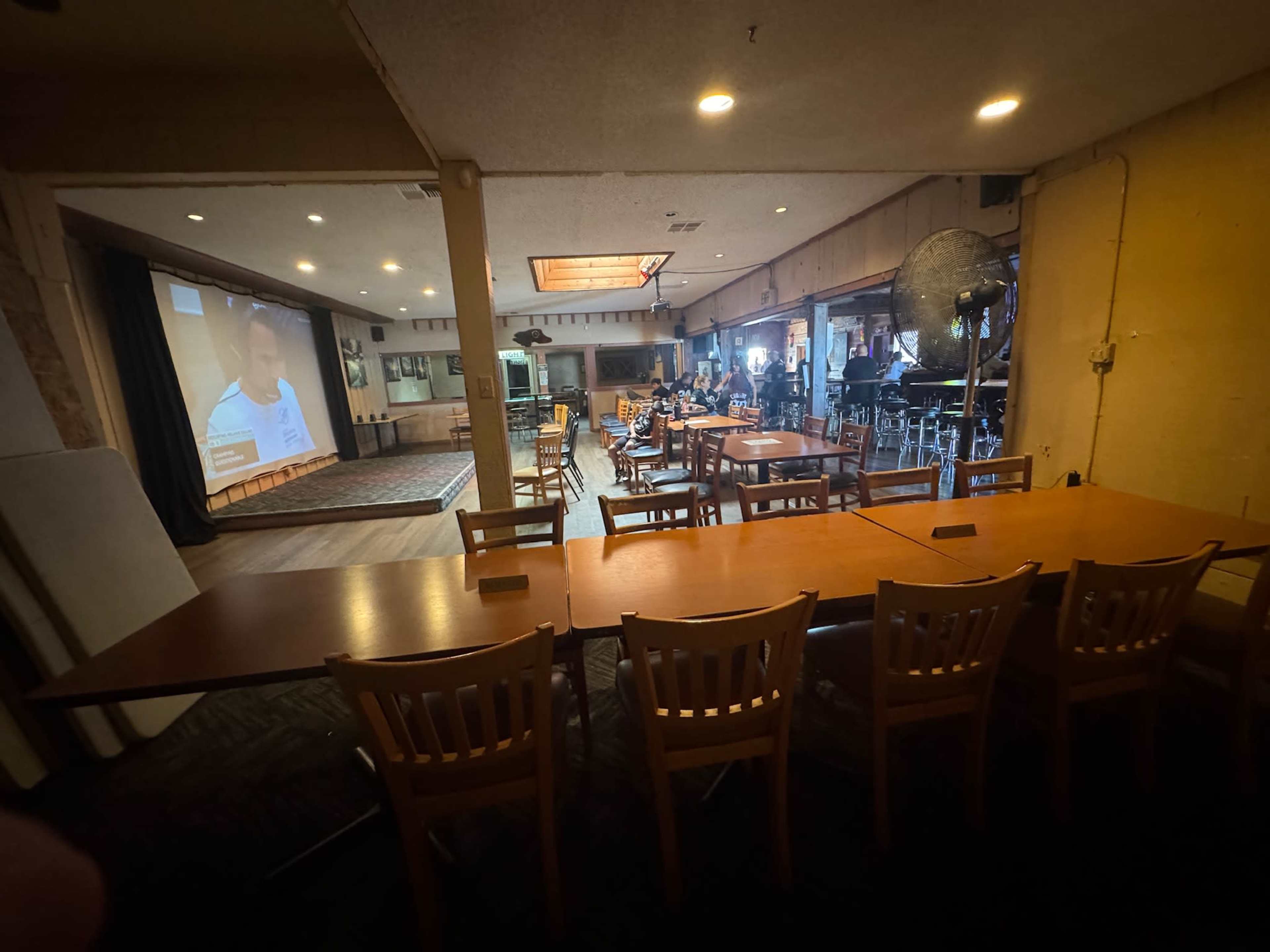 A long wooden table is set up in the foreground facing a large screen, while a group of people is seated at tables in a dimly lit room with a bar area in the background.