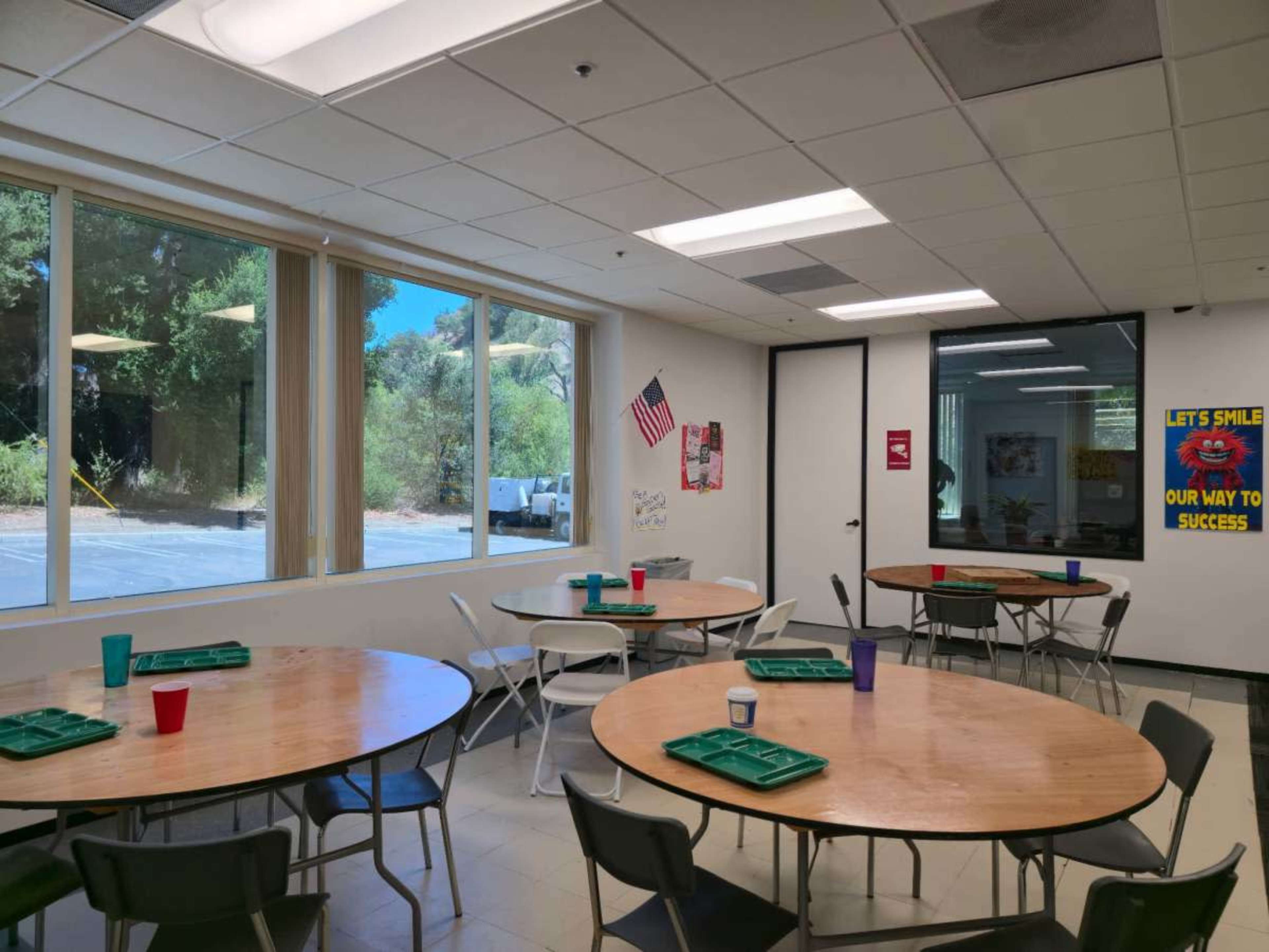 The image shows a dining area with multiple round tables and chairs, large windows letting in natural light, and a poster on the wall featuring an American flag and motivational messages.