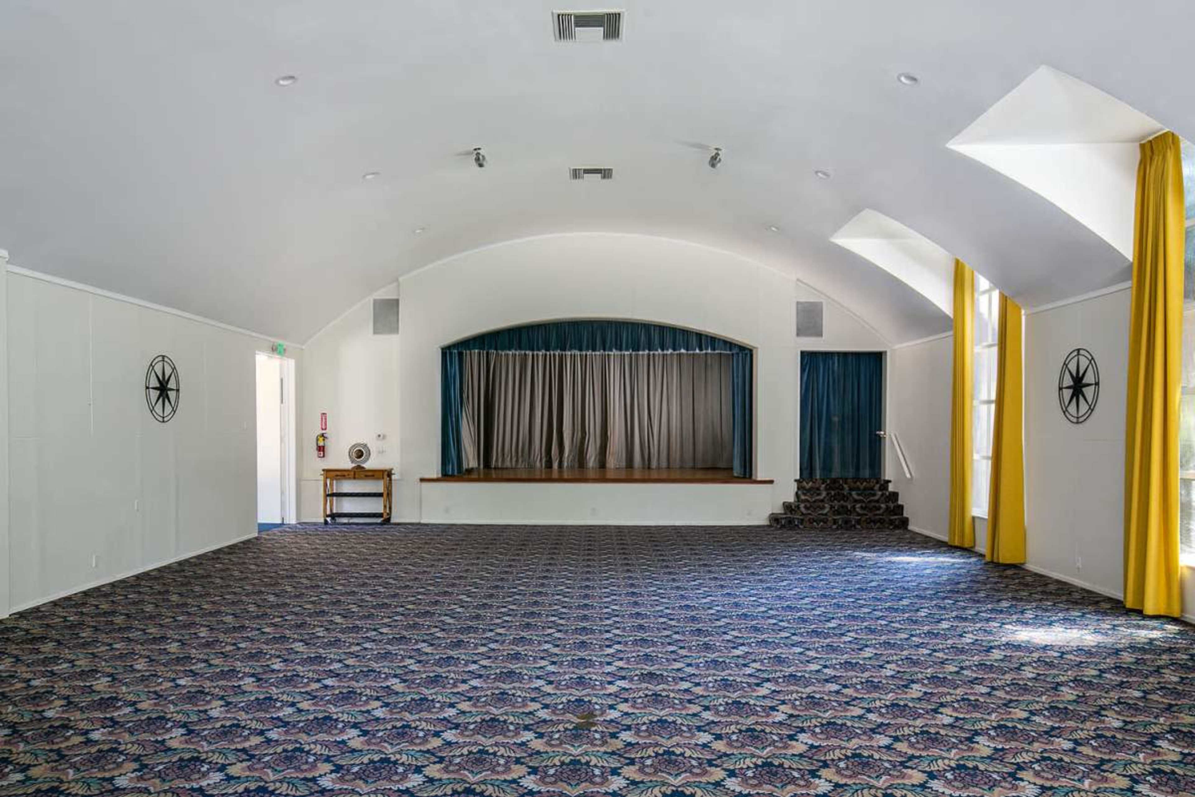 The image shows an empty room with a carpeted floor, a raised stage at the back, and large windows dressed with yellow curtains.