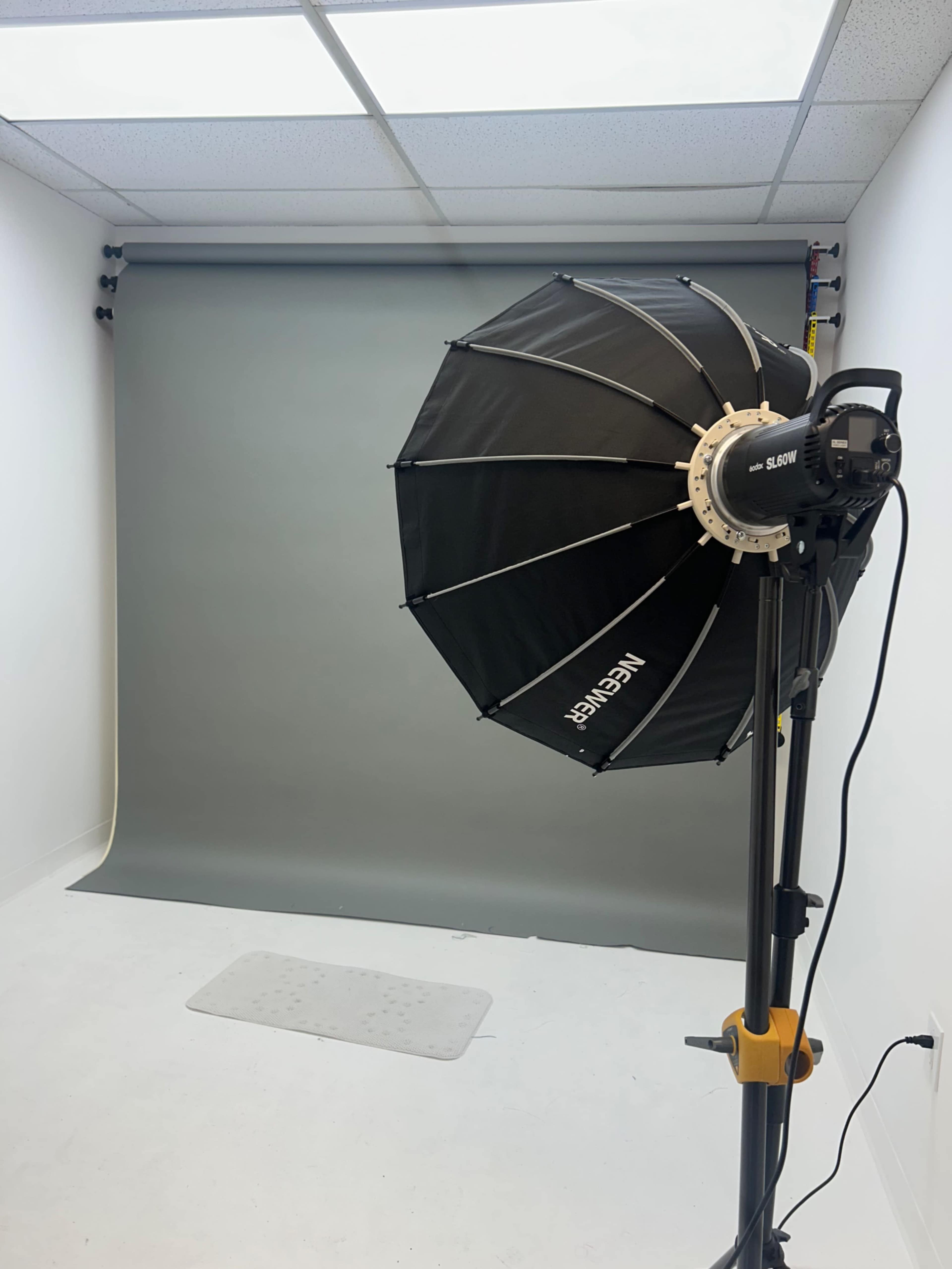 The image shows a photography studio equipped with a softbox light, a gray backdrop, and a clean, white floor.