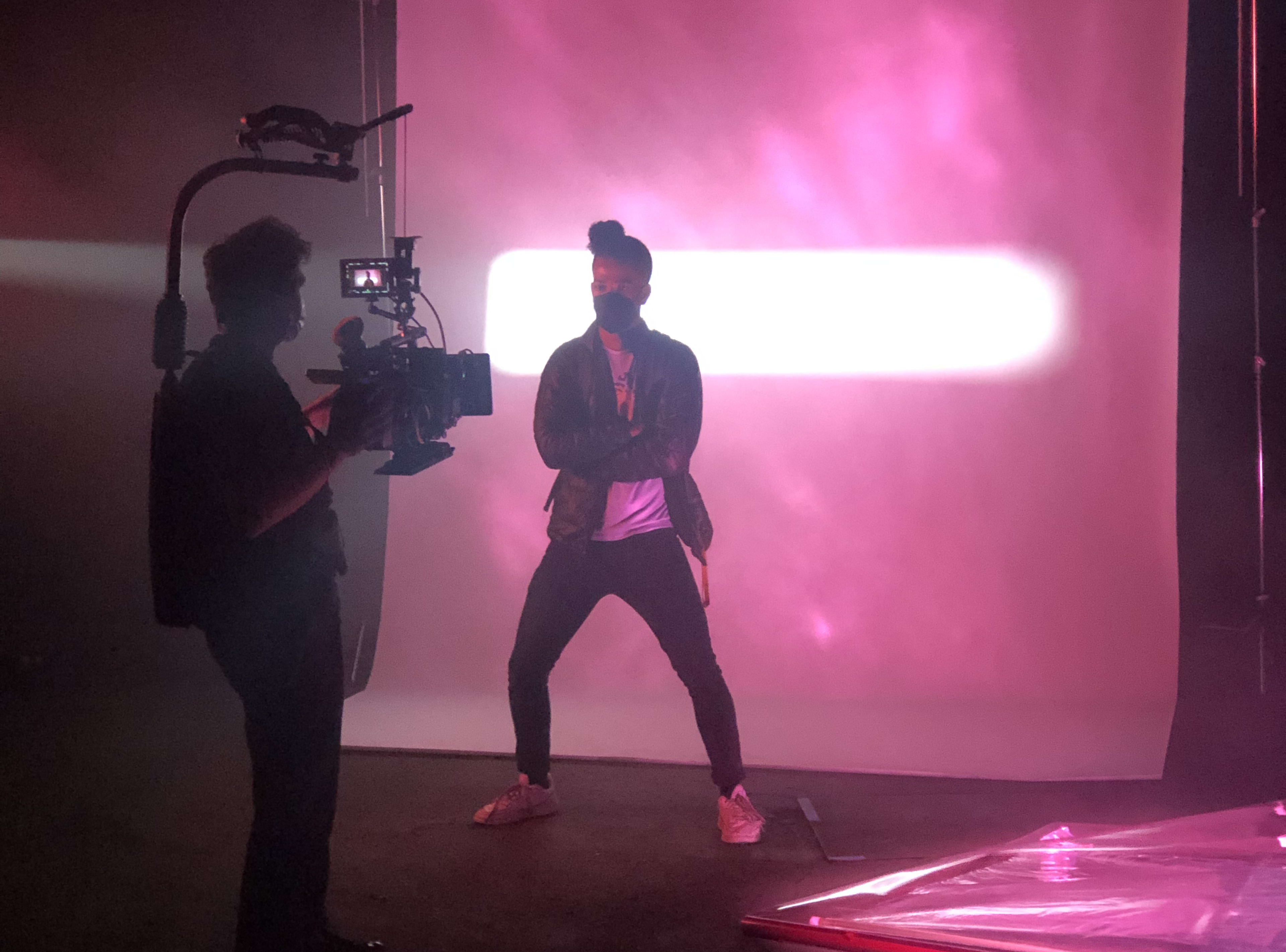 A person wearing a leather jacket and mask poses in front of a lit backdrop while another person films with a camera.
