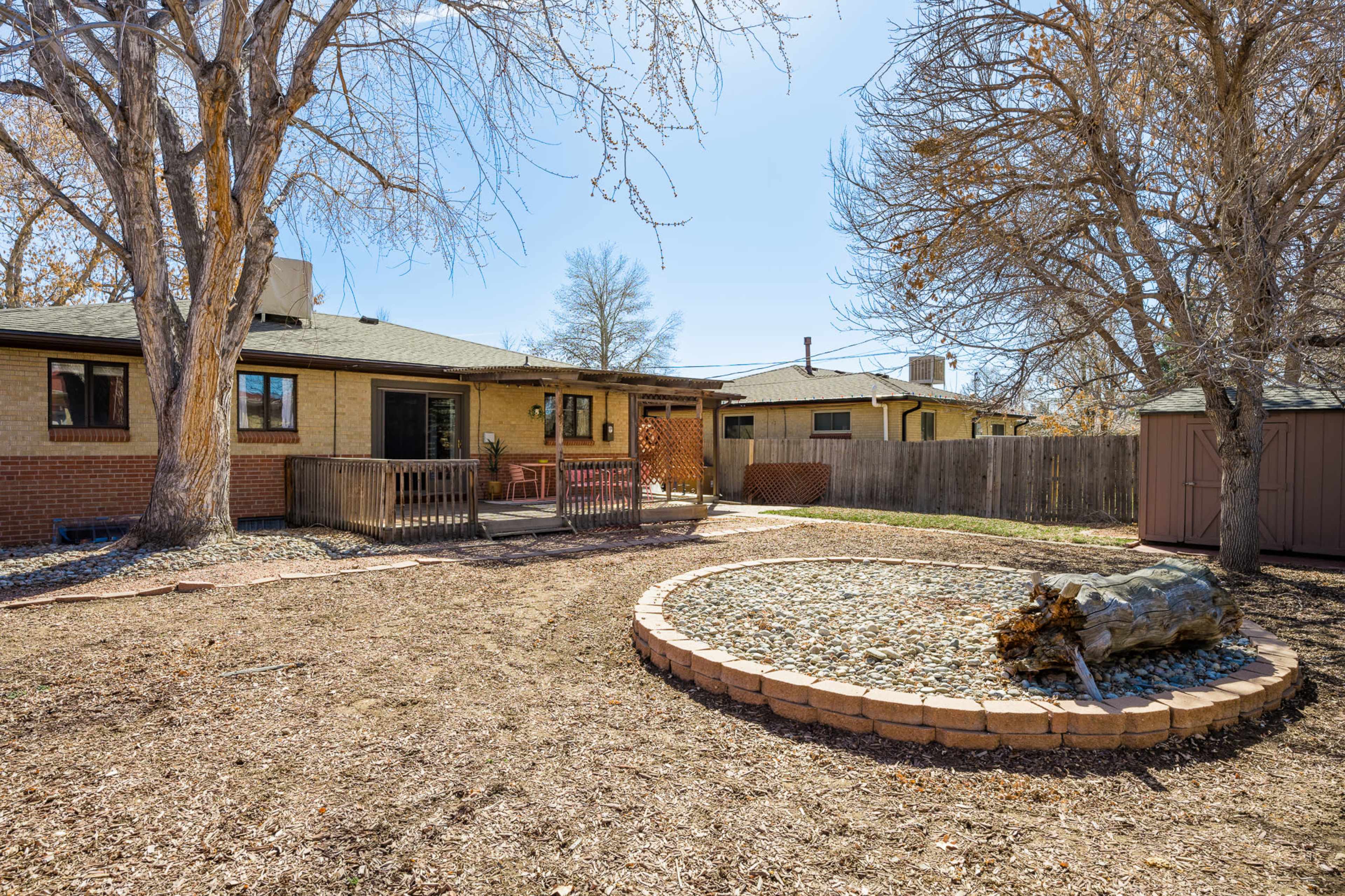 The image shows a backyard with a circular stone garden feature, a wooden deck, and trees, situated adjacent to a single-story home.
