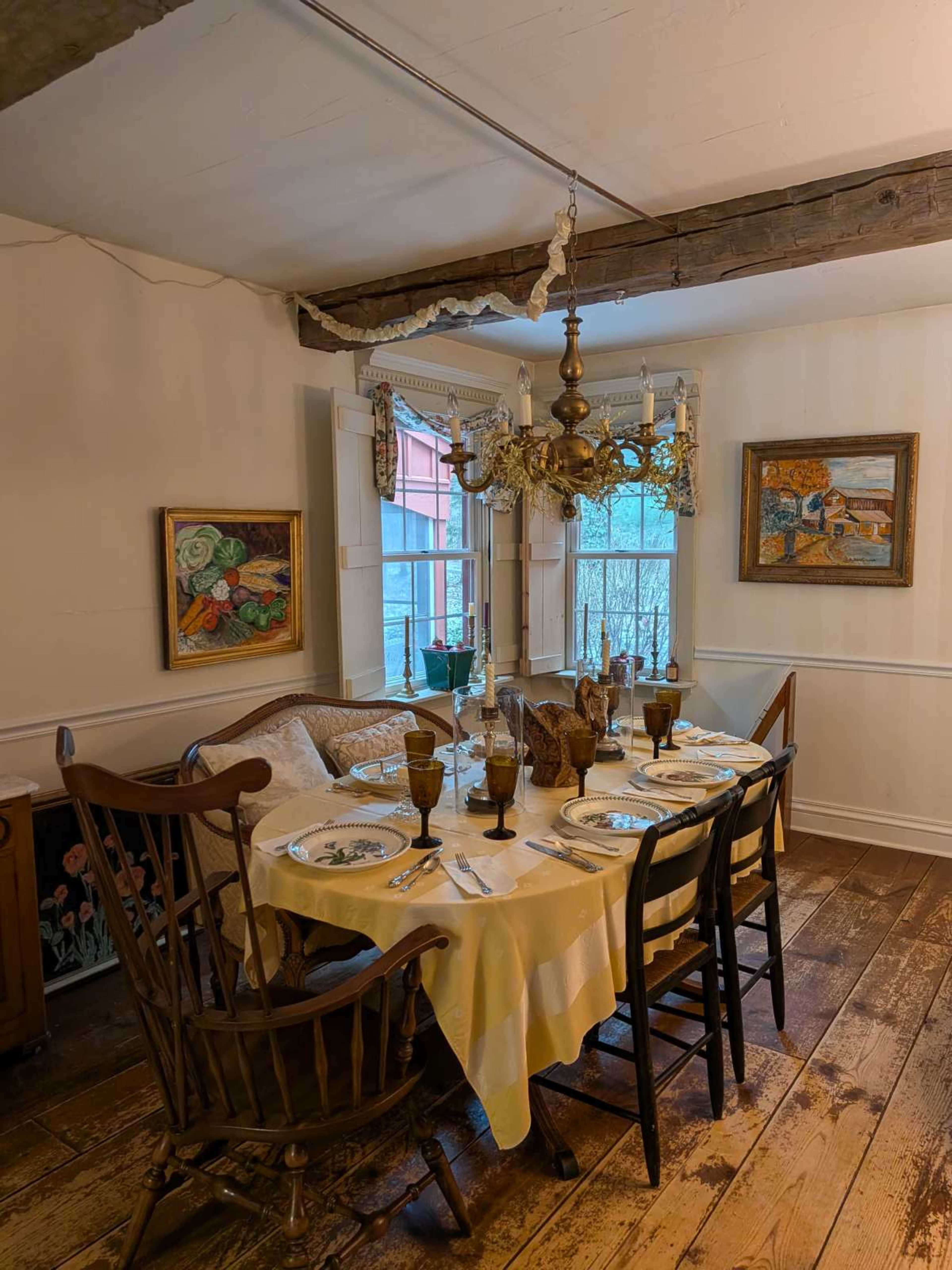 The image shows a dining area with a wooden table set for a meal, surrounded by chairs and adorned with decorative paintings and a chandelier.
