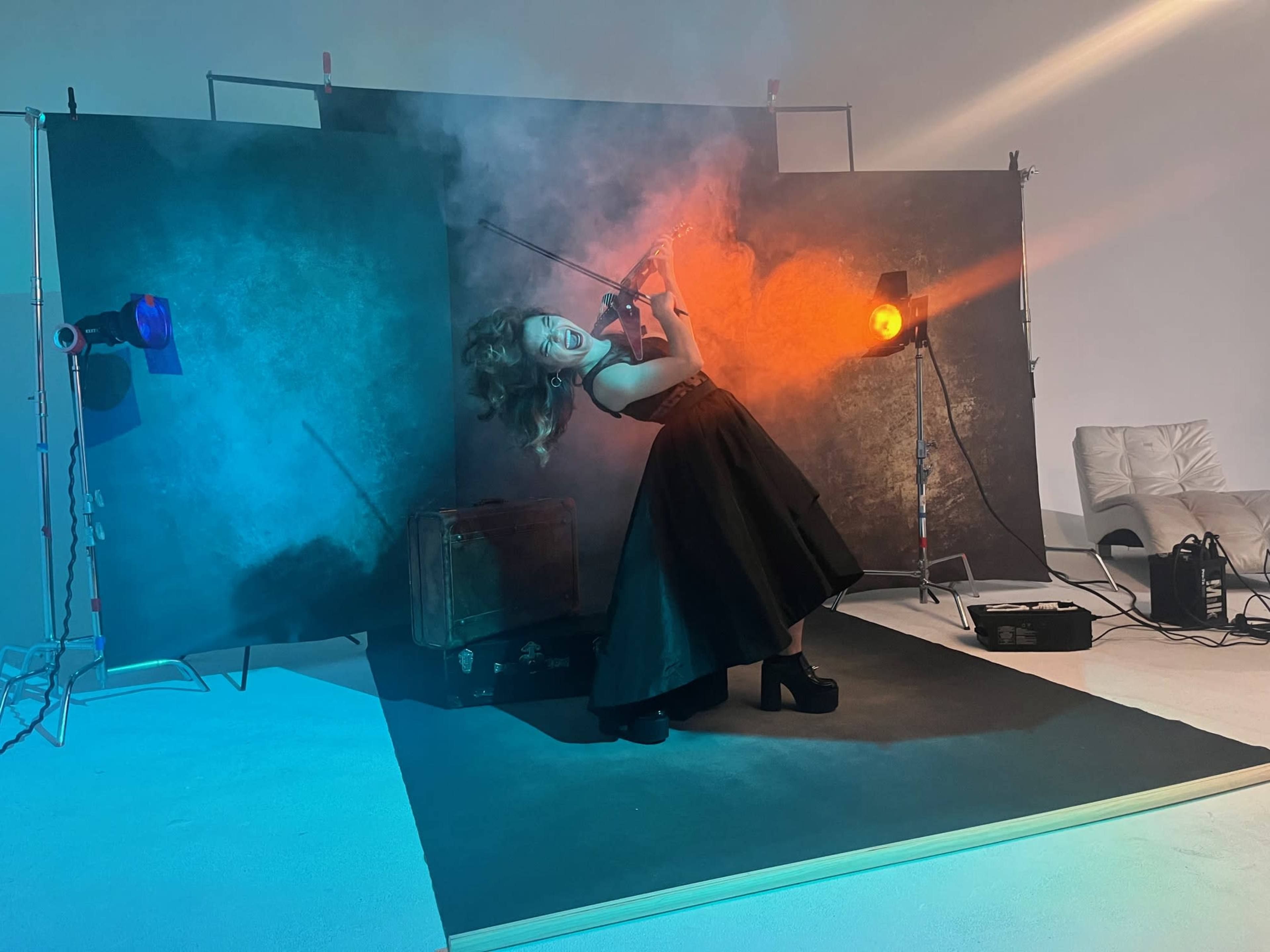 A woman in a black dress poses dramatically with a violin amidst colorful lighting and fog in a styled photo shoot setting.