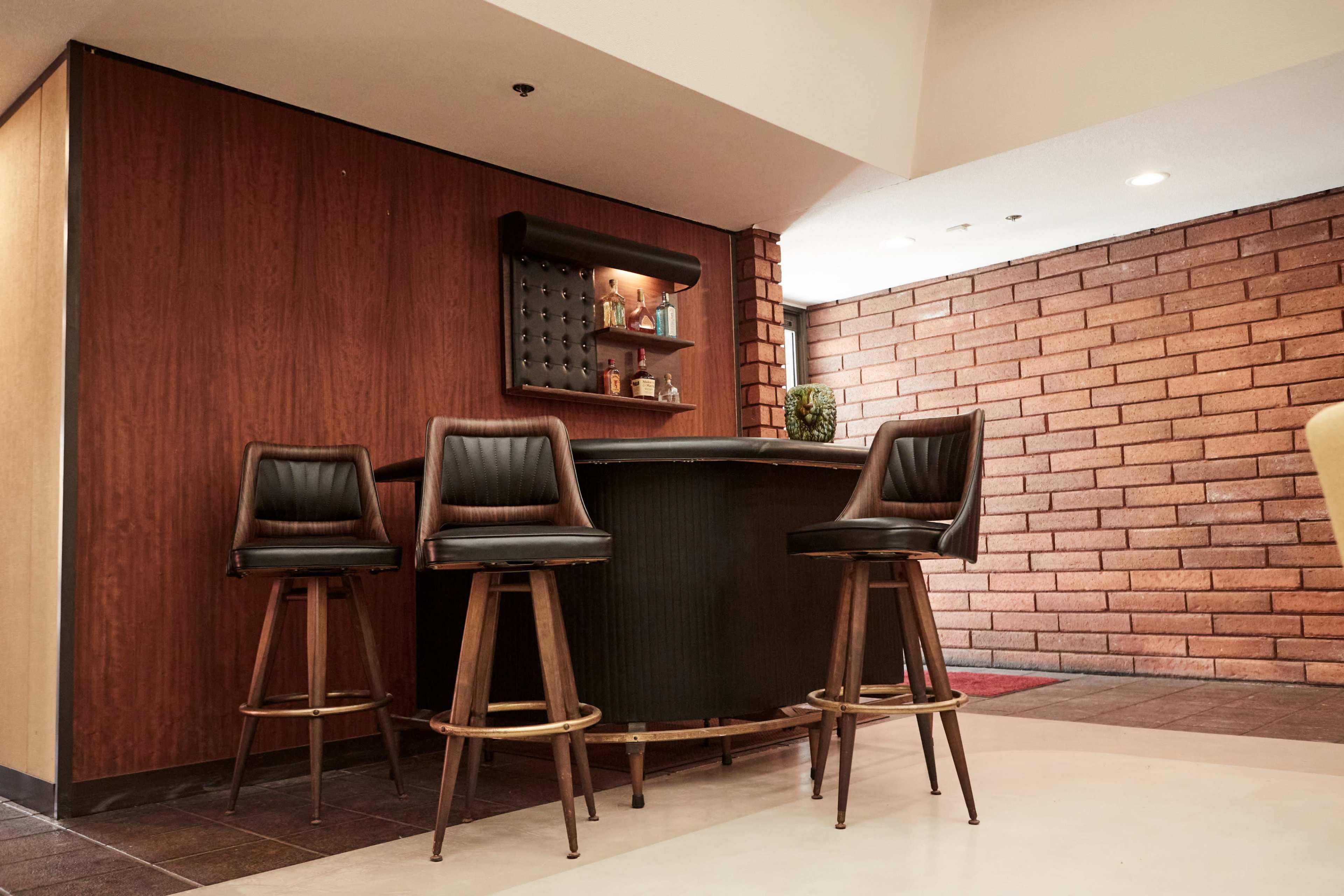 A small bar area with three black leather stools beside a circular table against a wooden wall with a shelf displaying decorative items.