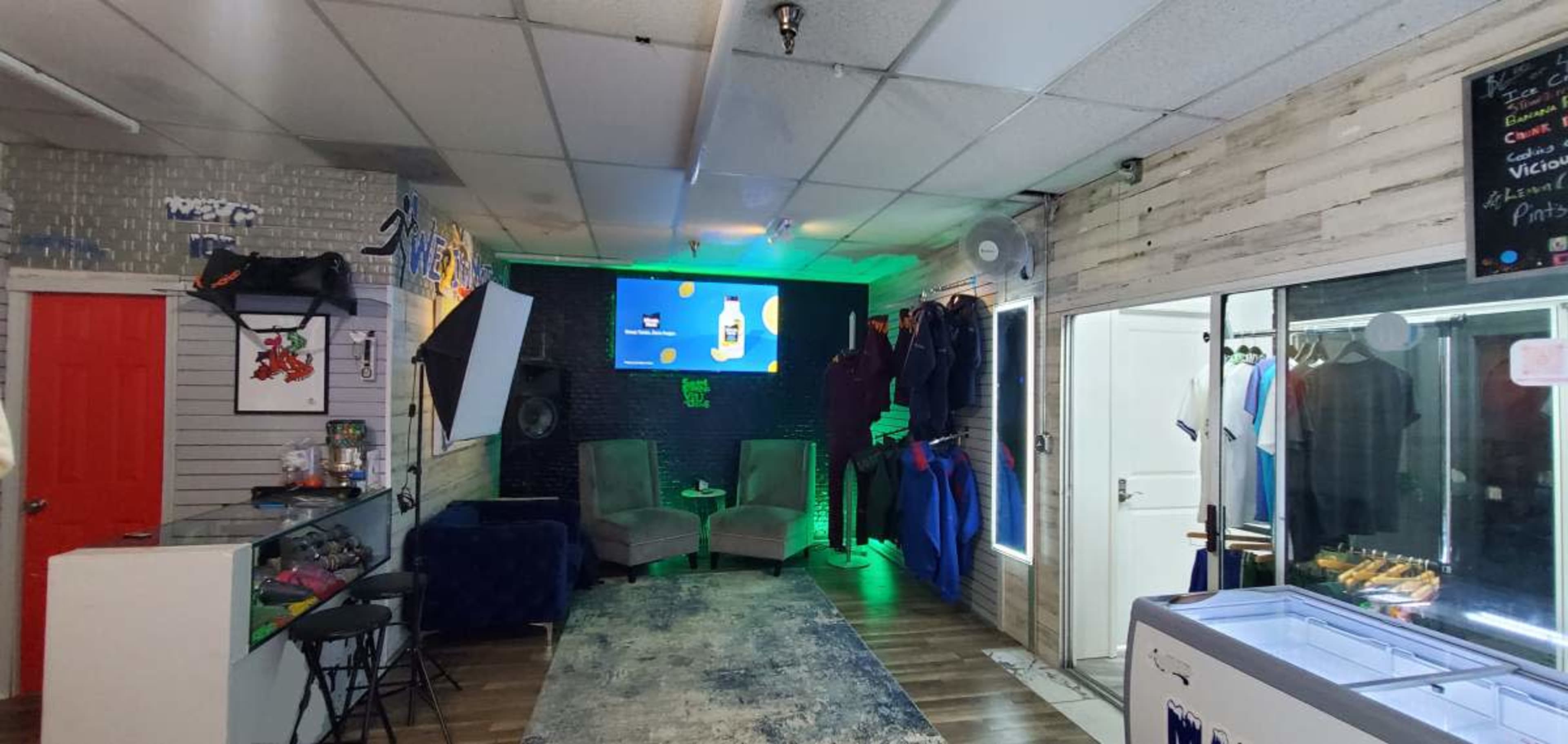 The image shows a neatly arranged interior space featuring two gray armchairs, a large television screen on the wall, and a glass display area filled with clothing items.