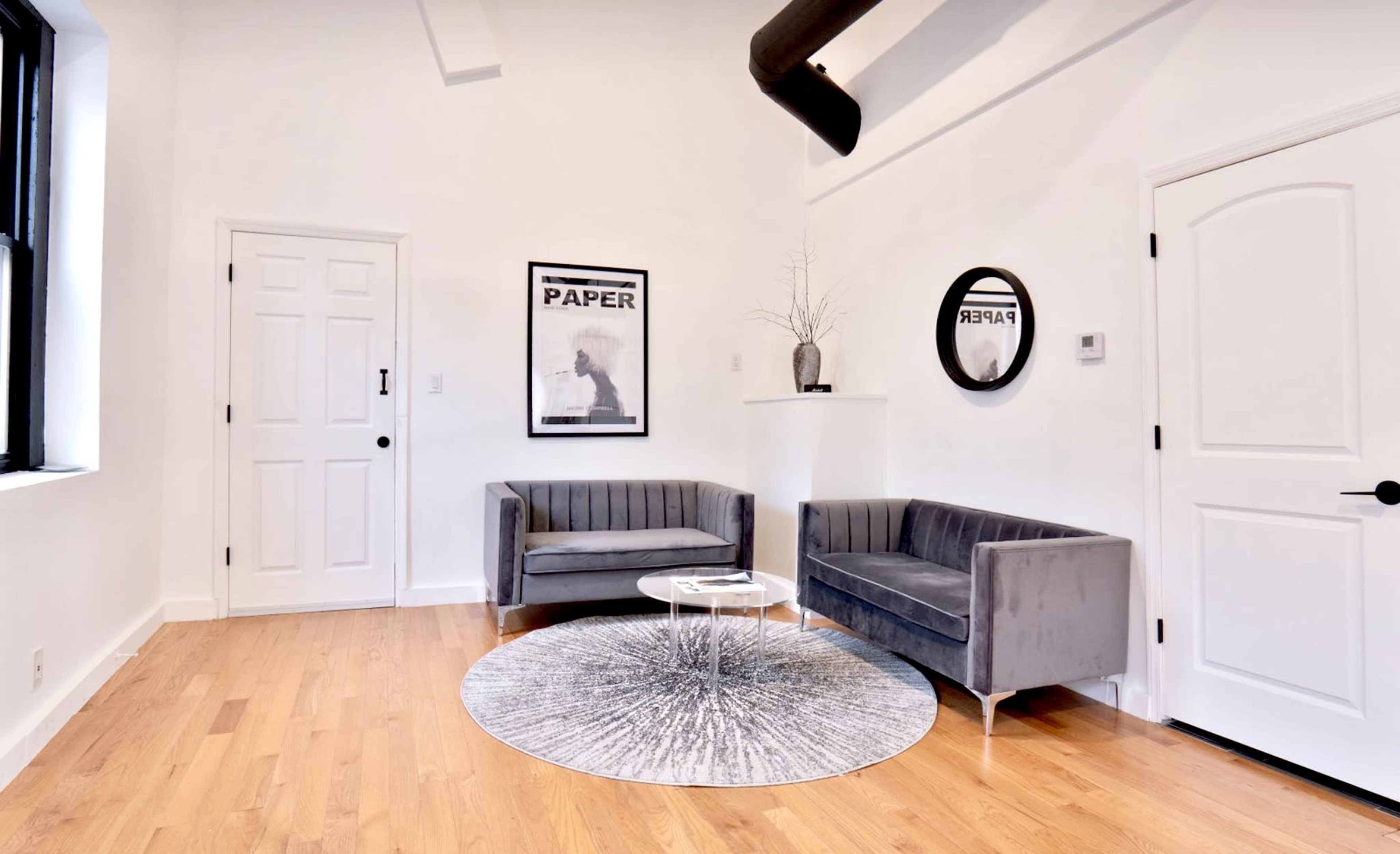 The image features a modern living room with two gray sofas, a round area rug, a round mirror, and a framed poster on a white wall.