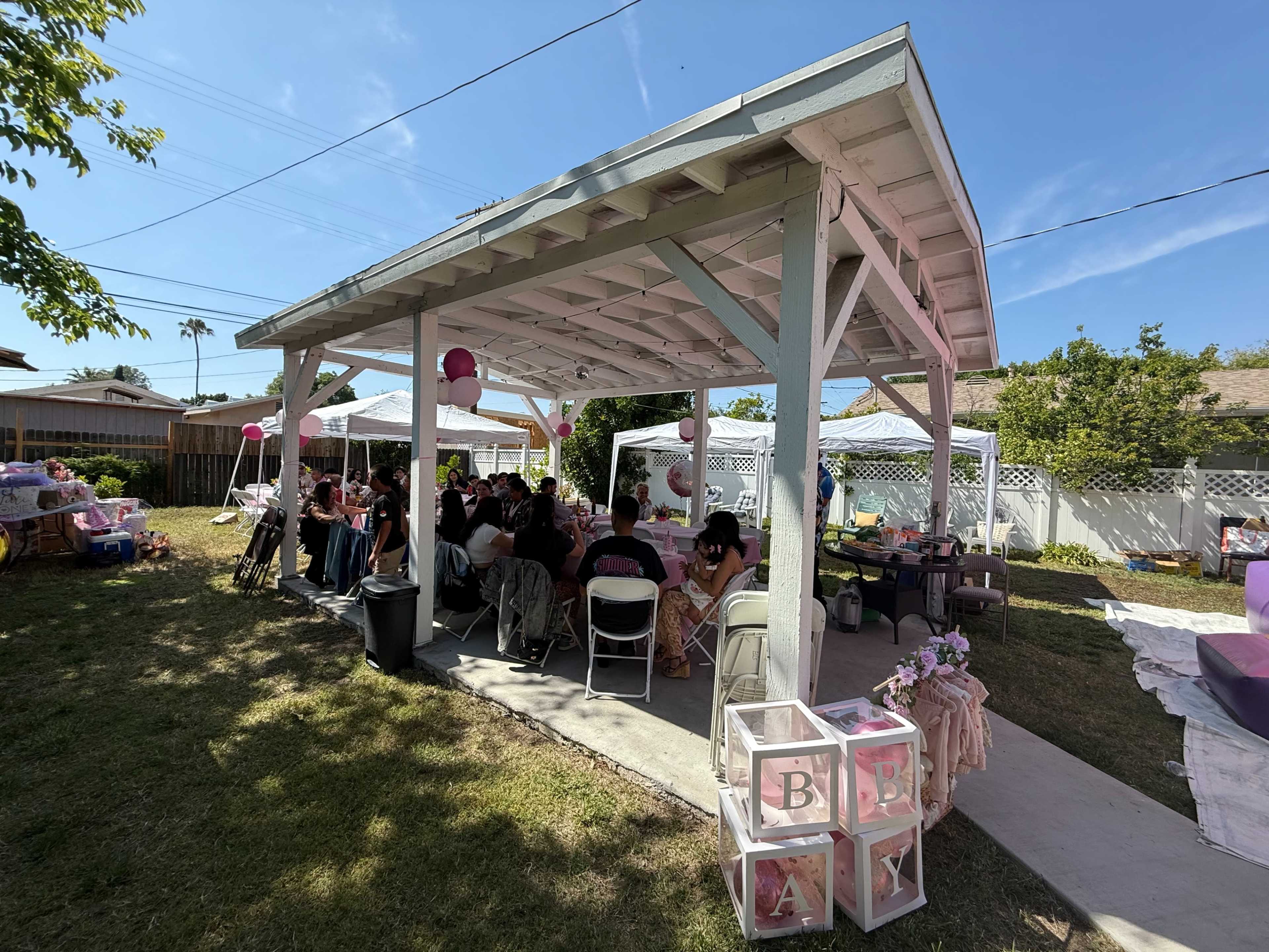 A gathering of people sits under a covered patio in a backyard, with decorated tables and party supplies visible.