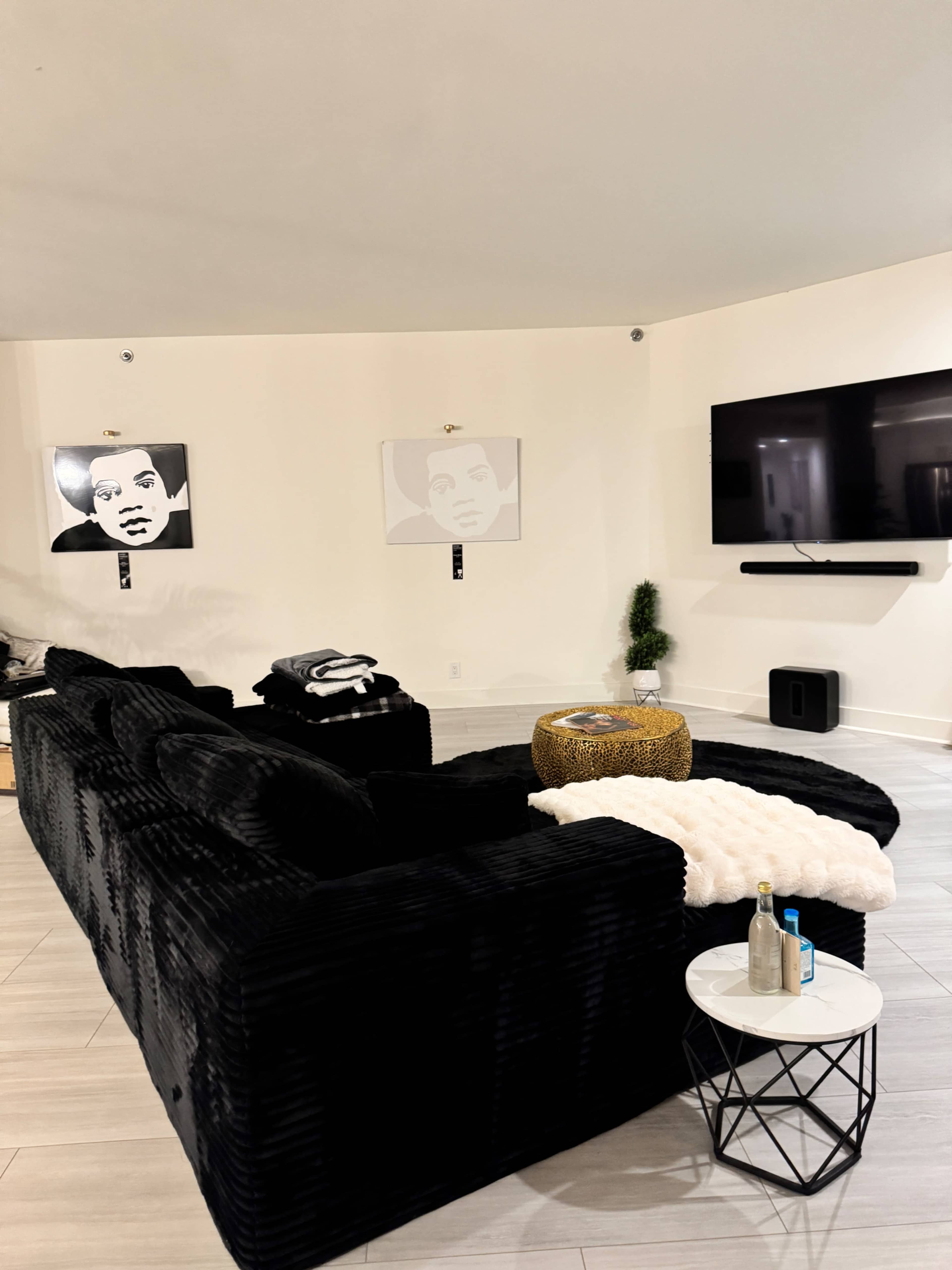 The image shows a modern living room with a black sectional sofa, a round coffee table, and two black-and-white portraits on the wall.