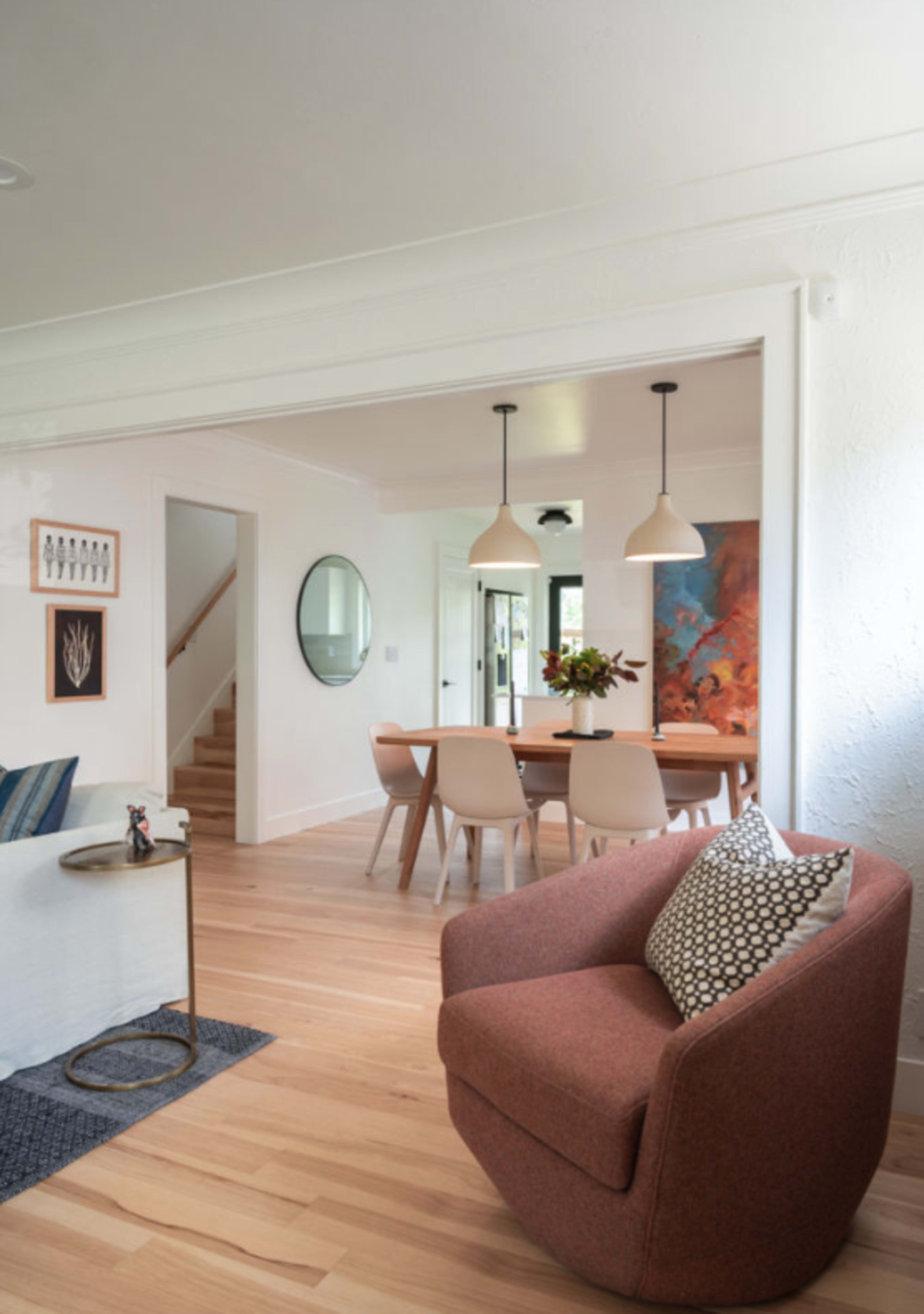 The image shows a modern living room open to a dining area, featuring a round dining table, stylish chairs, a staircase, and decorative artwork on the walls.