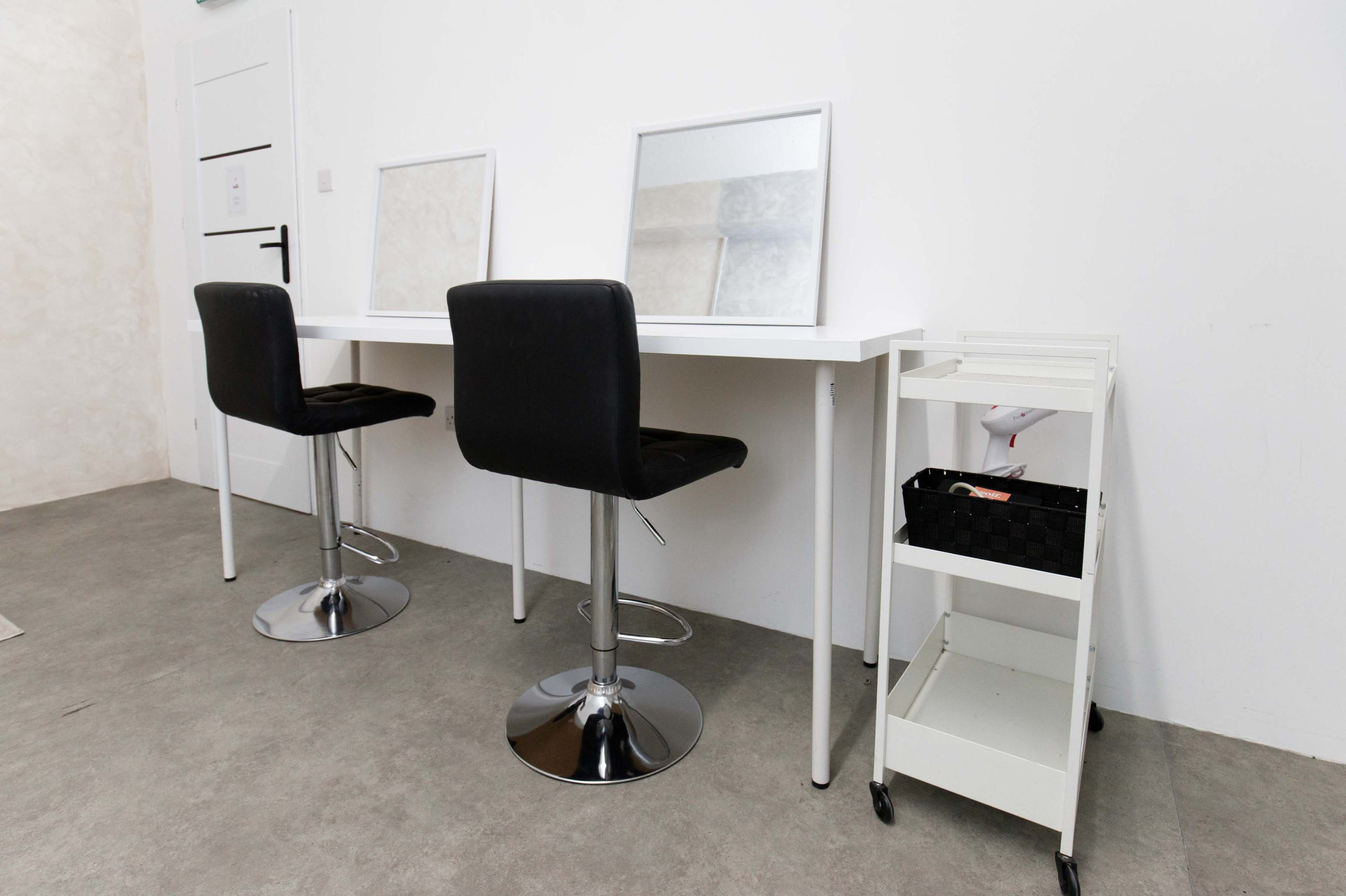 The image shows a minimalist makeup station with two high bar stools, a long white table, two mirrors, and a mobile storage cart beside it.