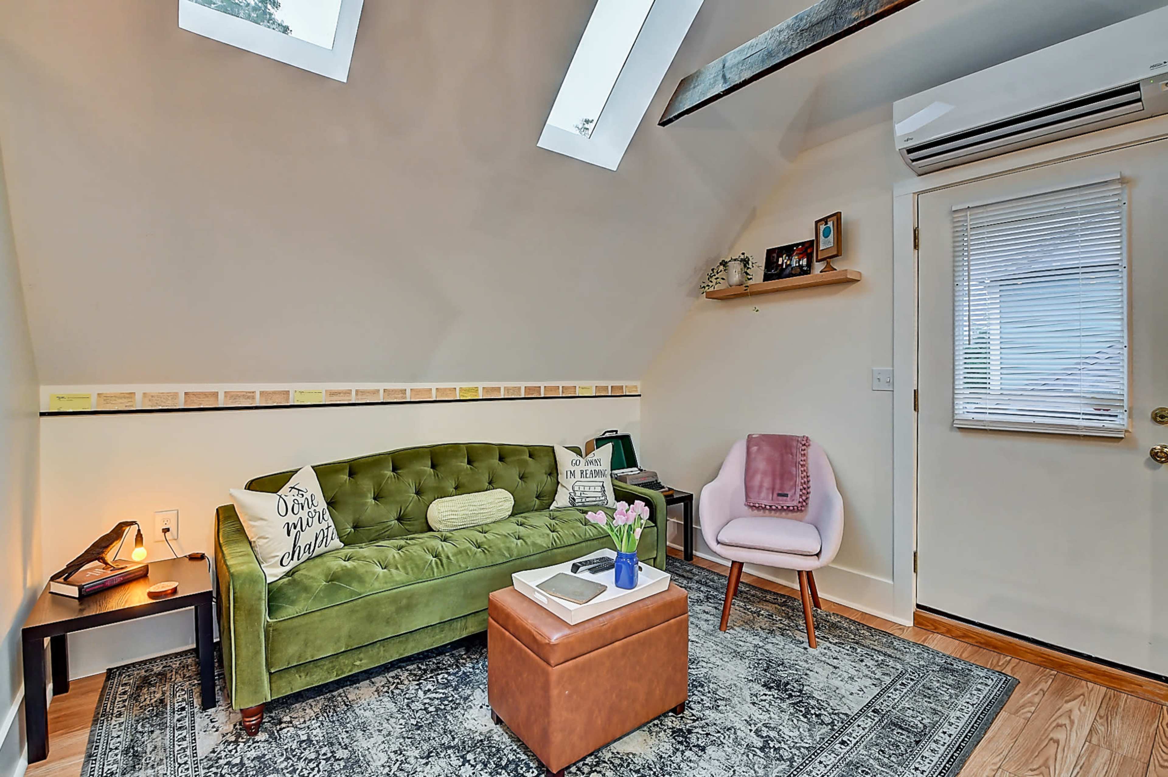 The image shows a small, cozy living area with a green velvet sofa, a pink chair, a wooden coffee table, and a decorative rug, illuminated by natural light from skylights.