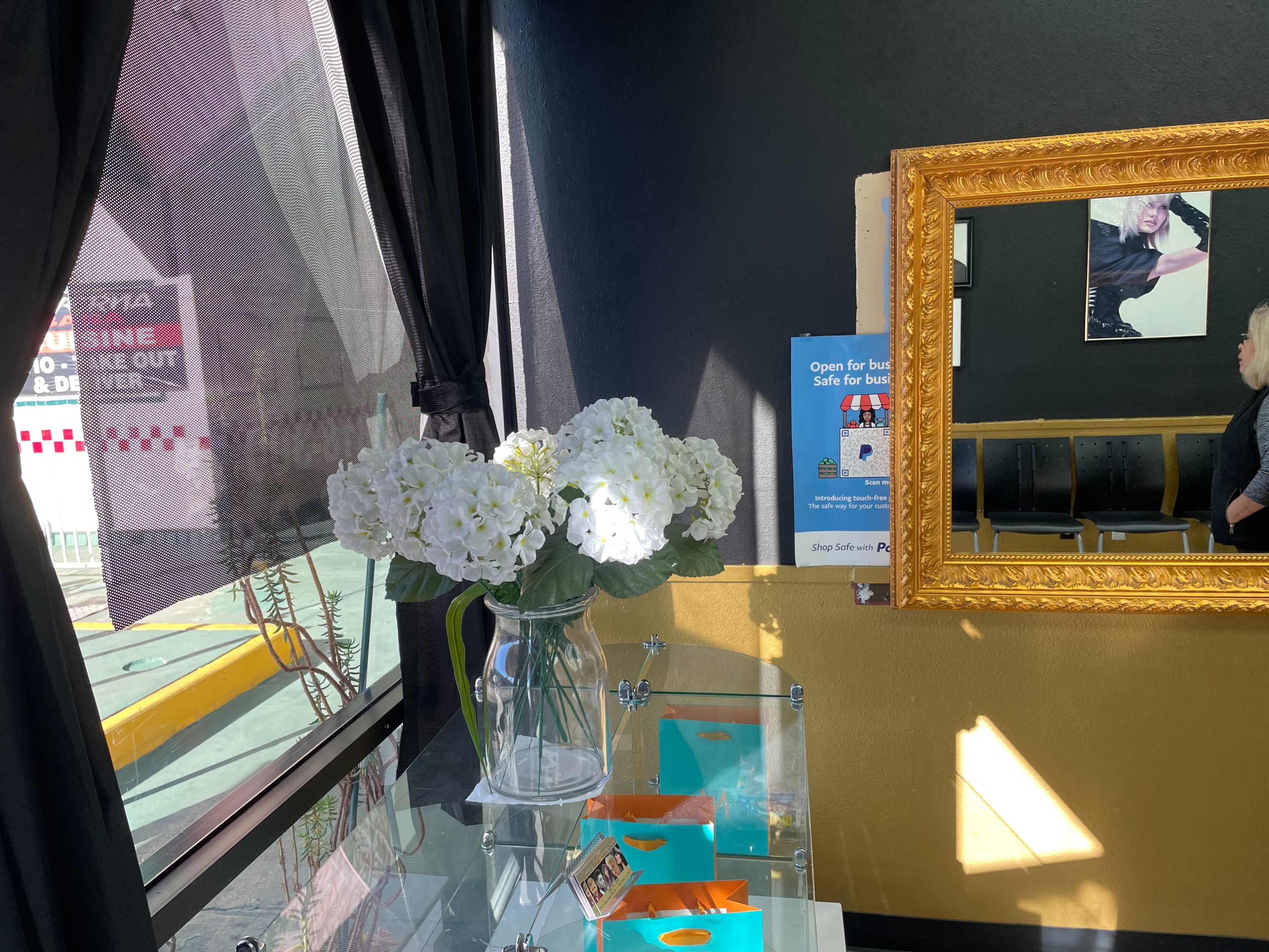 A vase of white flowers sits on a glass table next to a large framed mirror in a brightly lit indoor space.