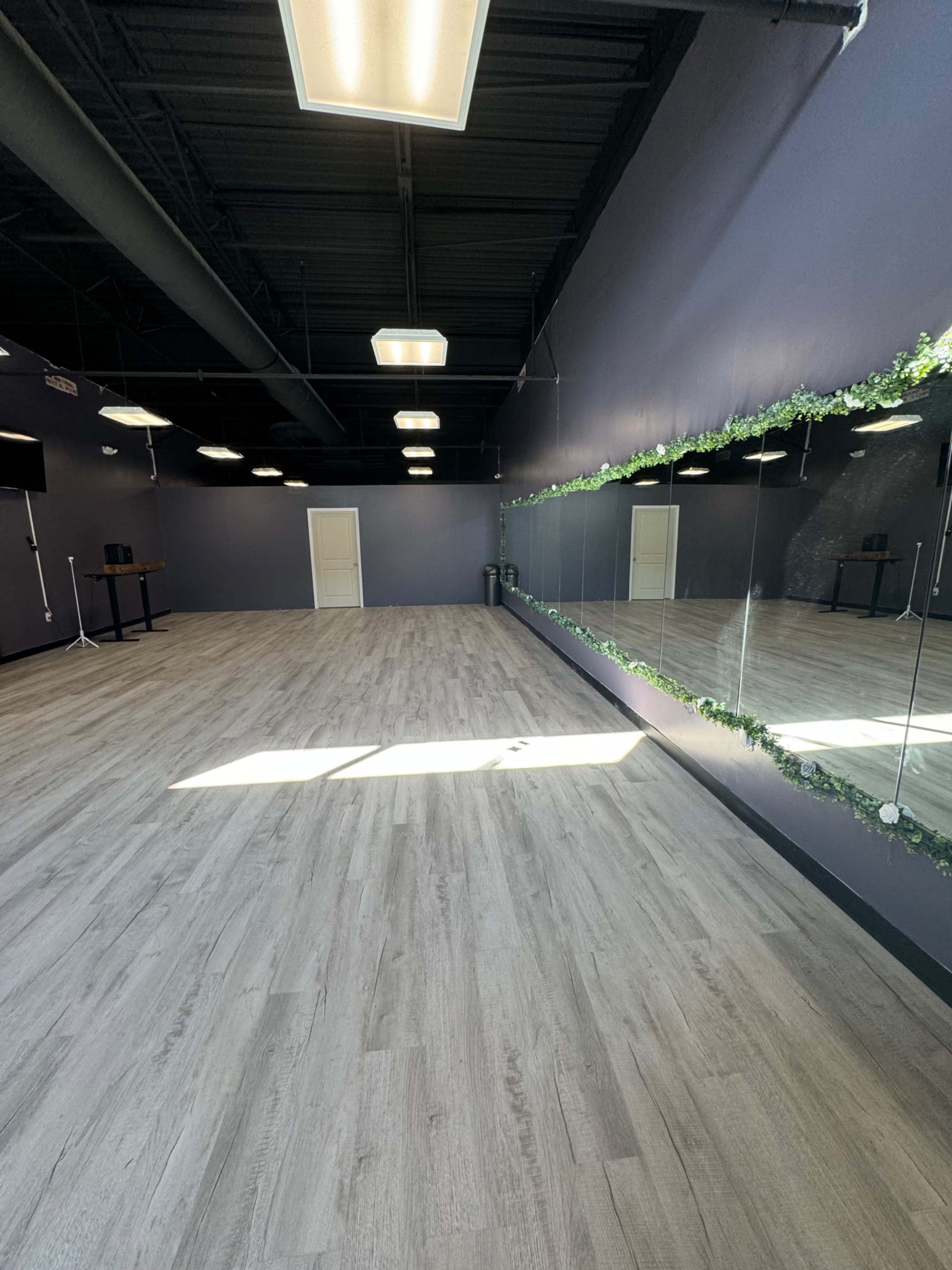 The image shows a spacious, empty room with wooden flooring and a large mirror on one wall, decorated with greenery.