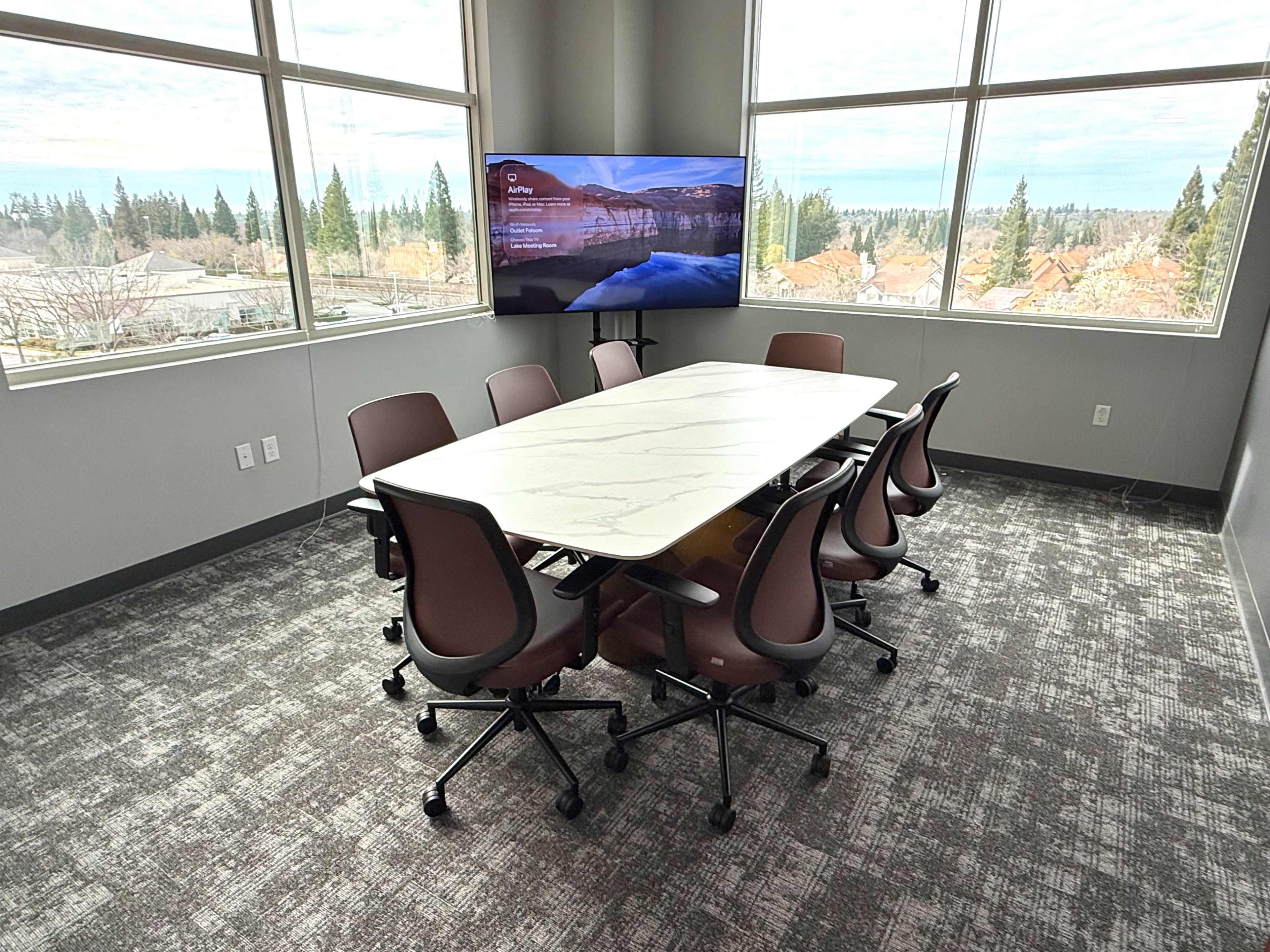 The image shows a modern conference room featuring a large marble-topped table surrounded by ergonomic chairs, with a screen displaying content on the wall and large windows overlooking a landscape.