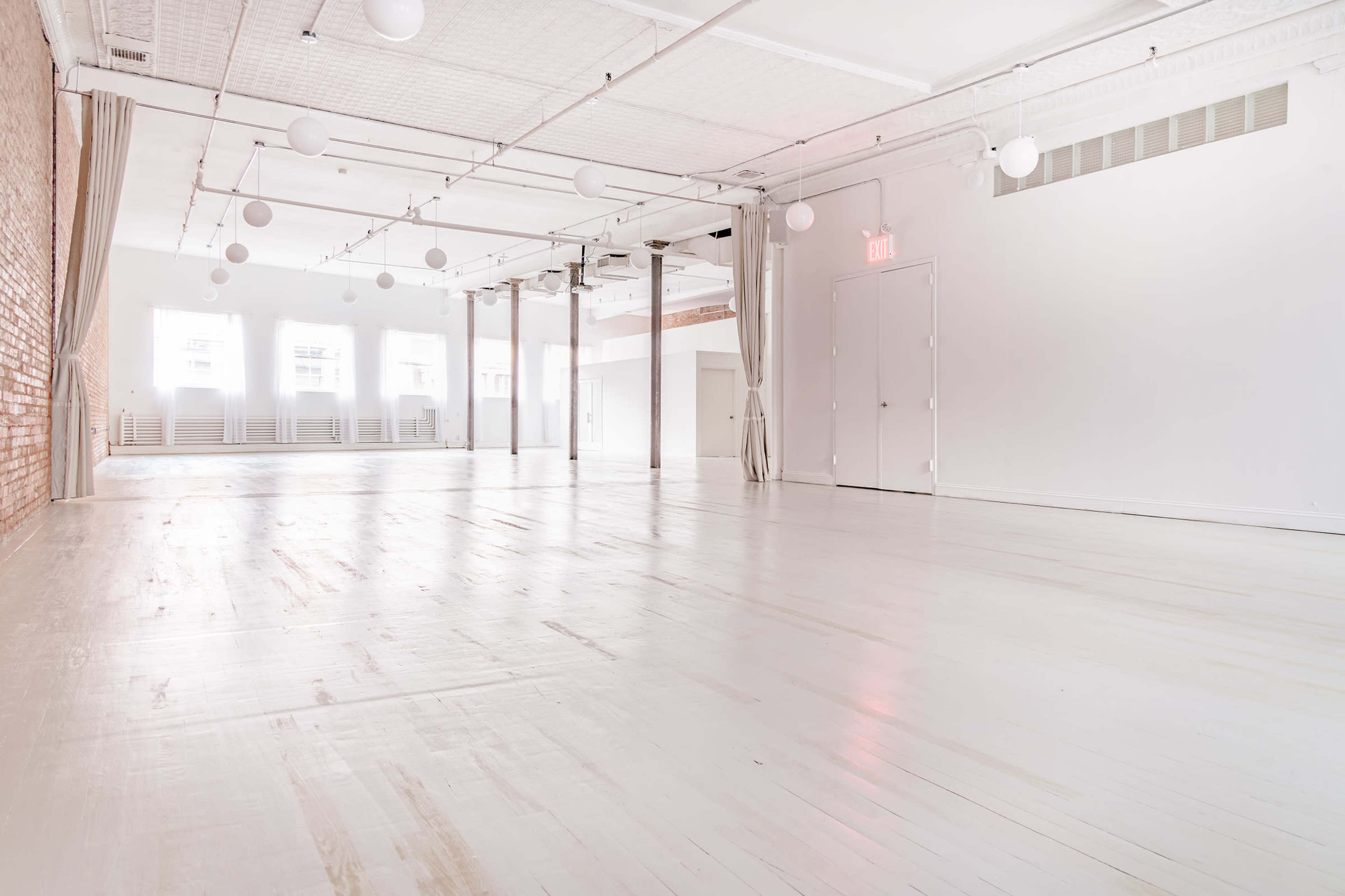 A spacious, brightly lit room with white walls and a polished wooden floor, featuring geometric light fixtures and some chairs against the back wall.