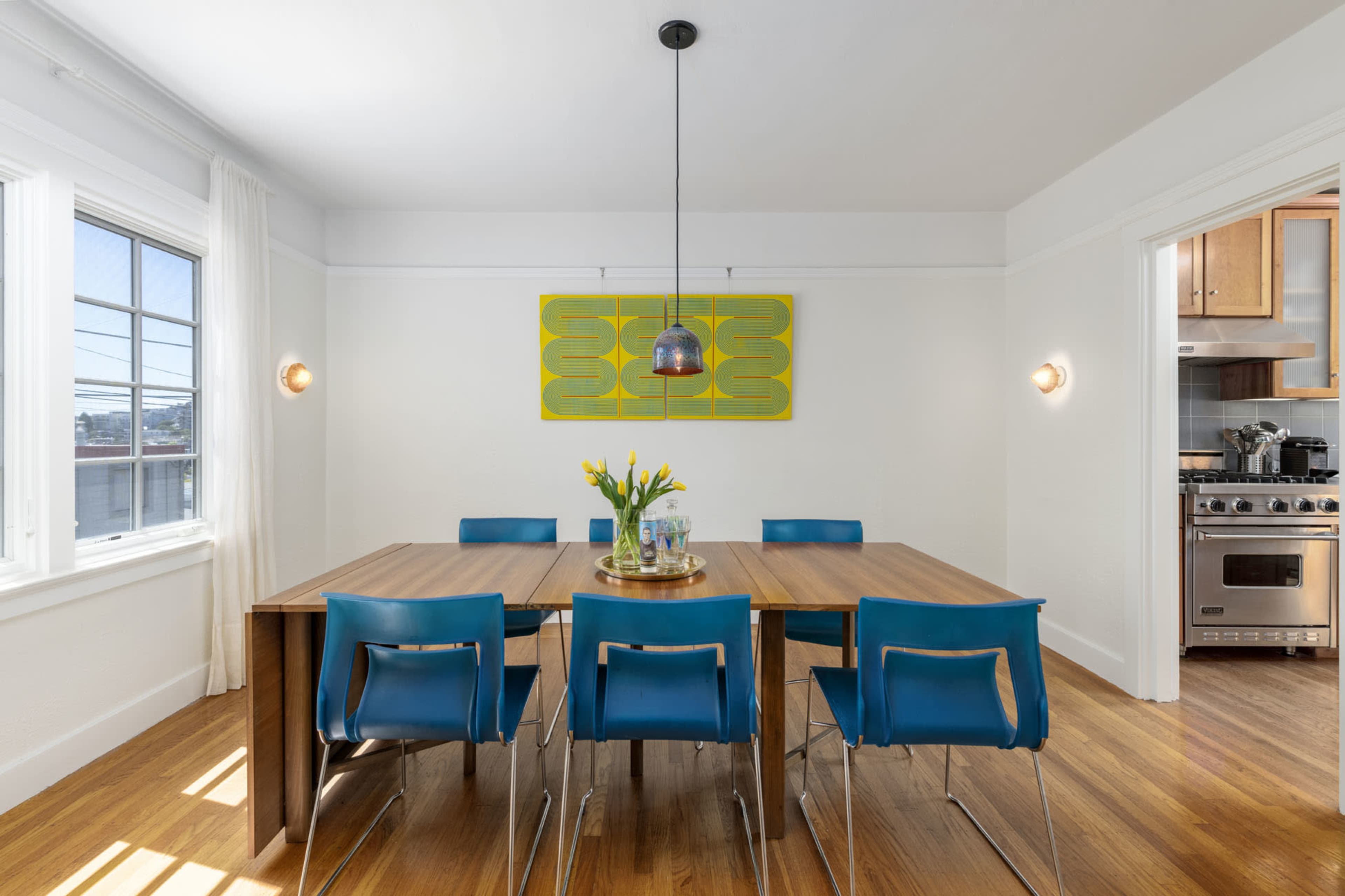 A dining room features a wooden table surrounded by blue chairs, with a yellow artwork on the wall and a pendant light above.