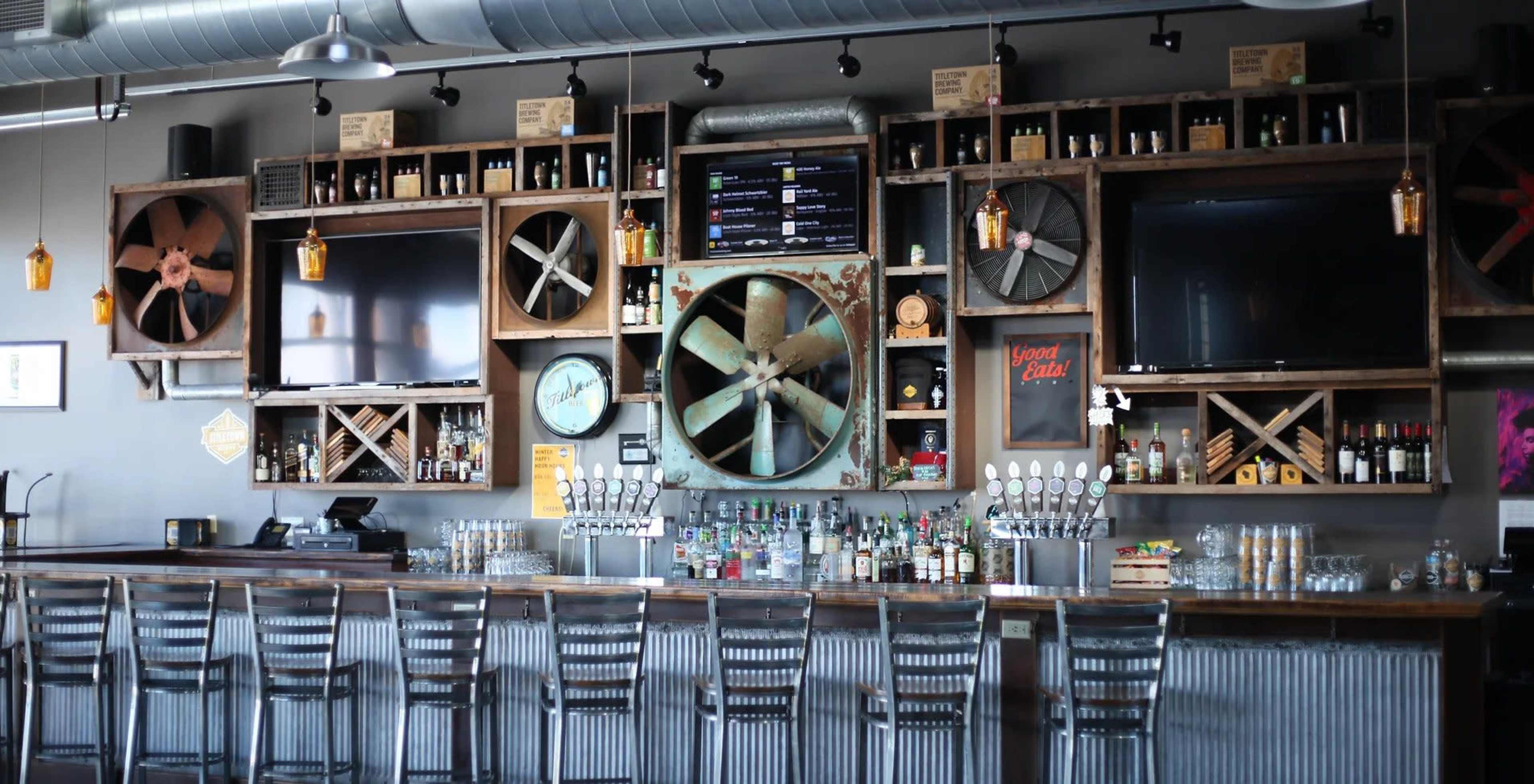 A trendy bar interior with a rustic wooden wall decorated with large fan replicas, televisions, and various bottles of liquor arranged on shelves.