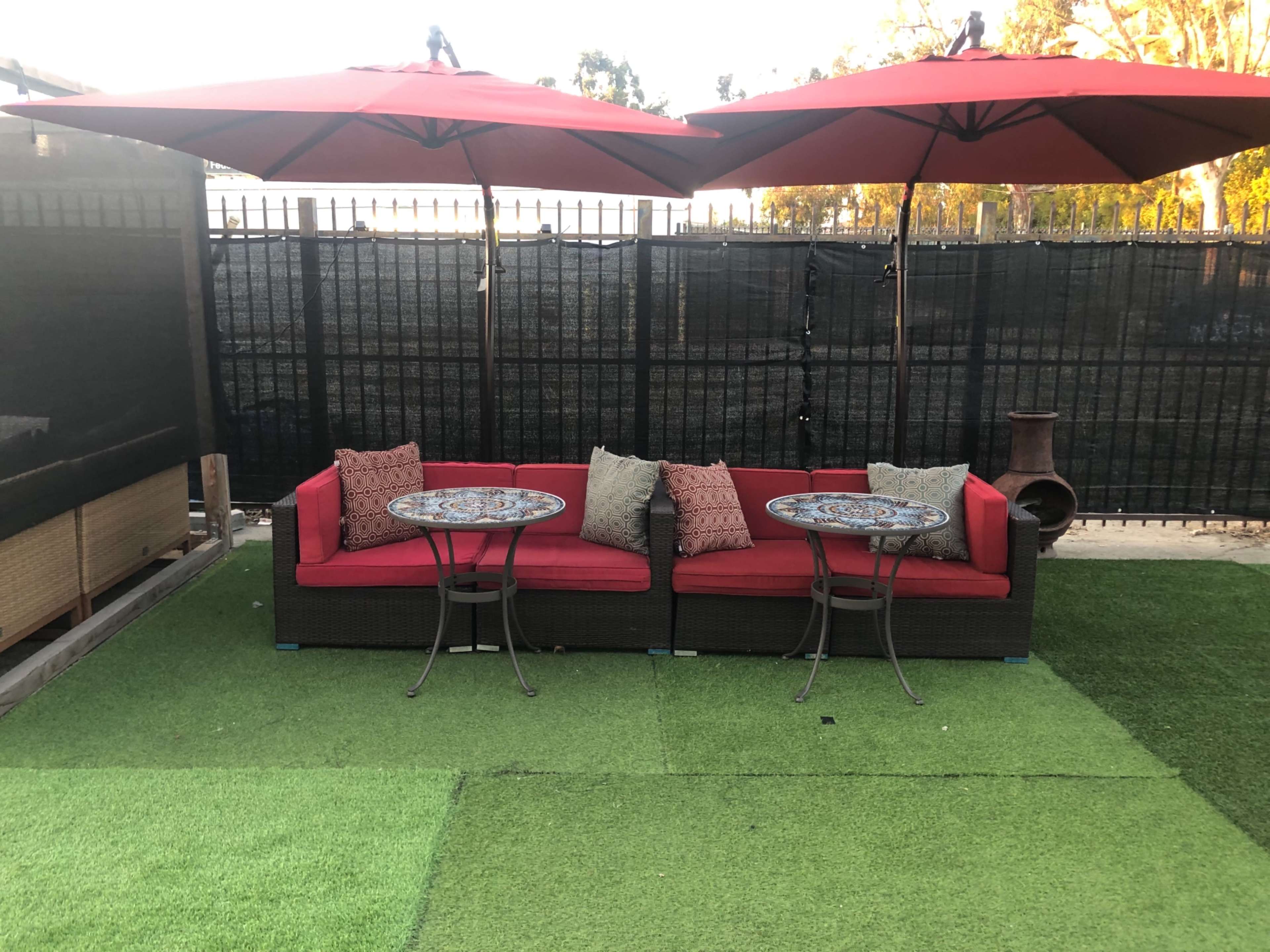 A seating area features two red umbrellas shielding a couch and two small round tables on artificial grass.