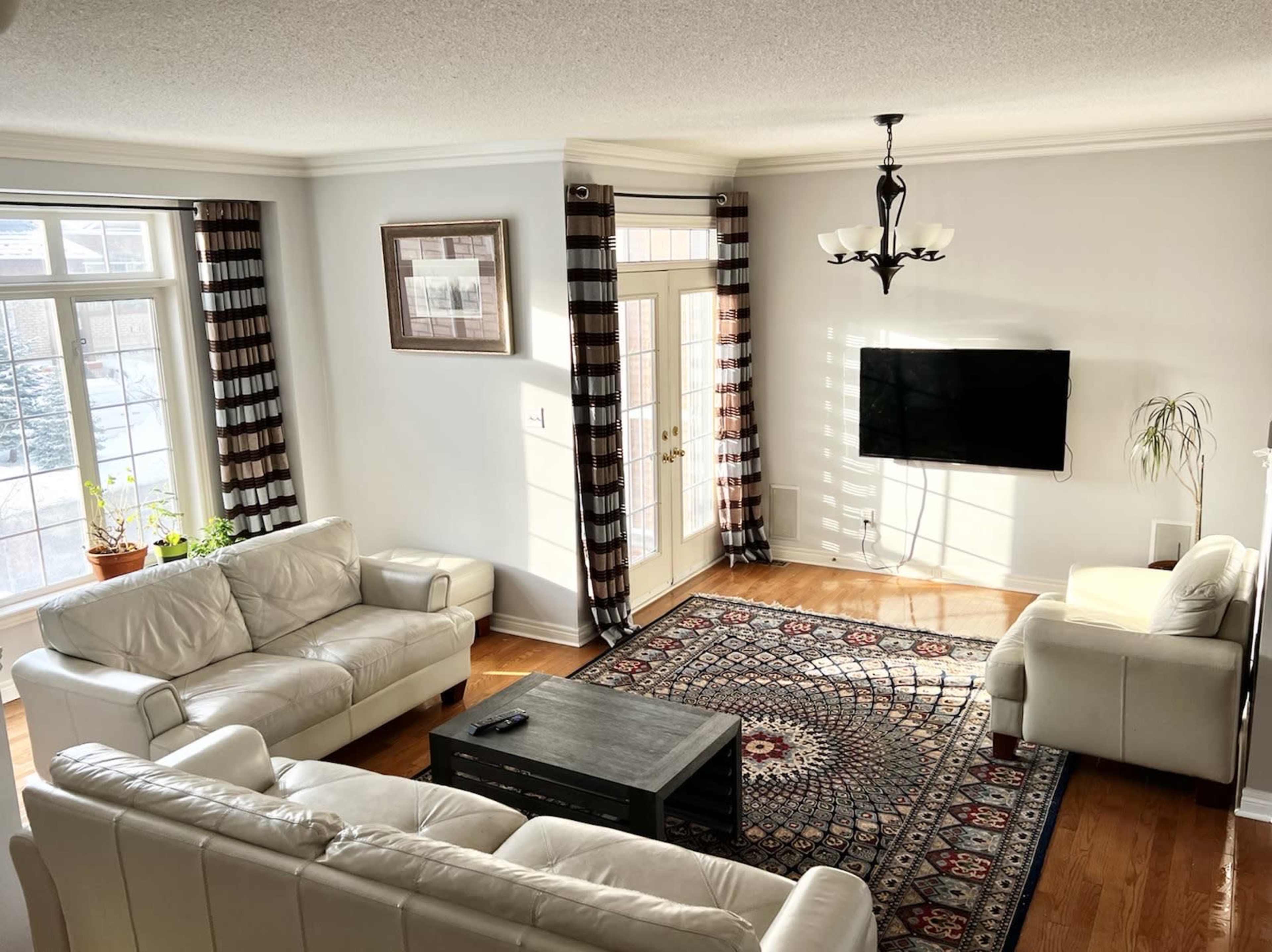 A living room features white leather sofas, a dark wooden coffee table, and large windows with striped curtains, along with a television mounted on the wall.